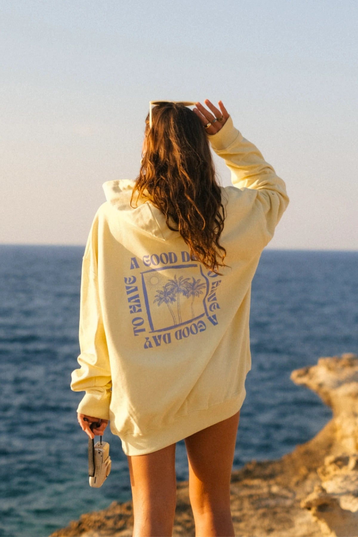 Frau trägt den A Good Day Oversized Hoodie in Butter mit großem Rückenprint von The Sunkissed Rebels an einer Steilküste mit Blick auf das Meer bei goldenem Sonnenlicht.