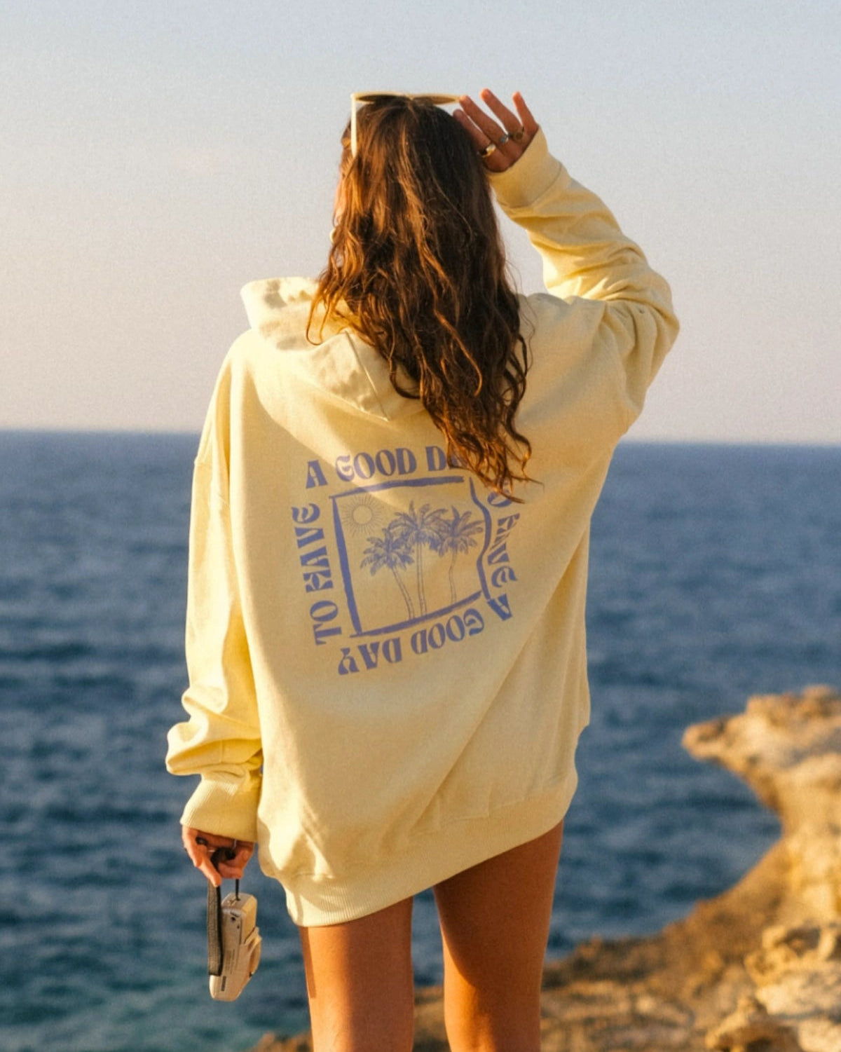 Frau trägt den A Good Day Oversized Hoodie in Butter mit großem Rückenprint von The Sunkissed Rebels an einer Steilküste mit Blick auf das Meer bei goldenem Sonnenlicht.