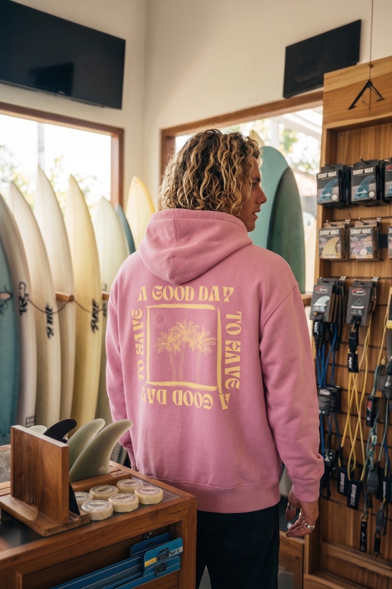 Rückenansicht des pinken A Good Day Hoodies mit großem Backprint in einem Surfshop mit Surfboards und Beach Lifestyle Atmosphäre