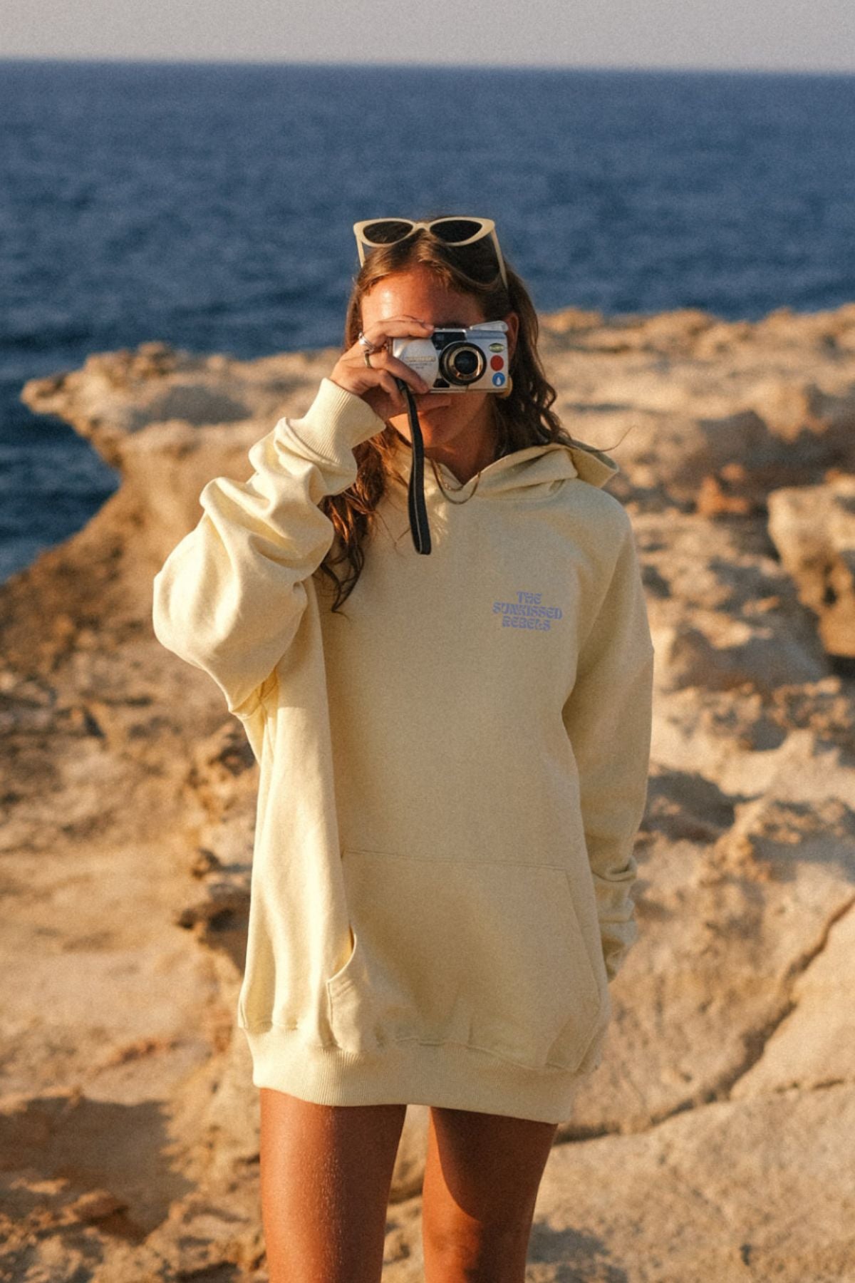 Frau trägt A Good Day Hoodie von The Sunkissed Rebels an den Klippen am Strand und macht ein Foto.