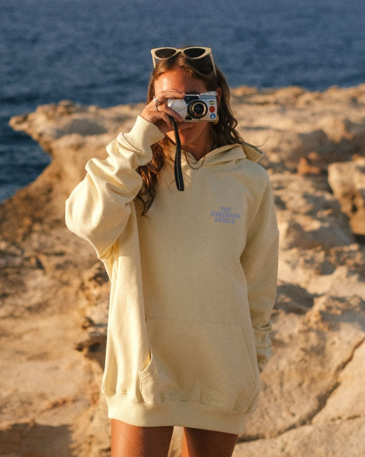 Frau trägt A Good Day Hoodie von The Sunkissed Rebels an den Klippen am Strand und macht ein Foto.