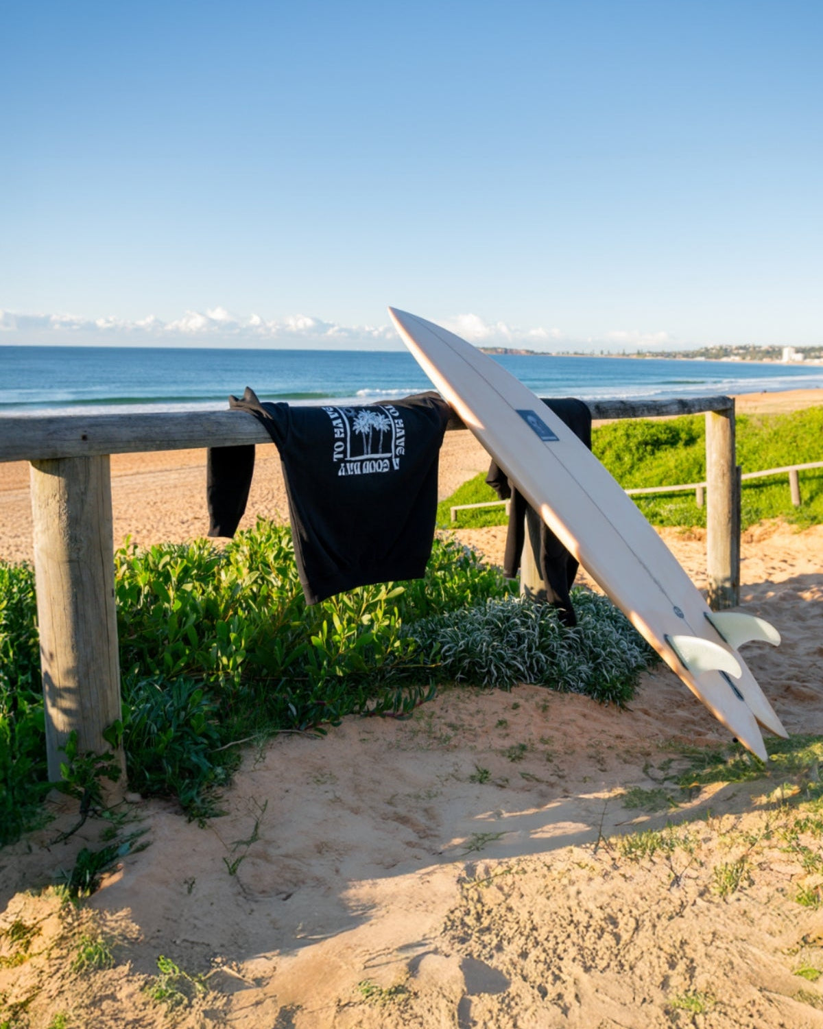 Der Sweater A Good Day von The Sunkissed Rebels hängt über einen Zaun am Strand neben einem Surfbrett.