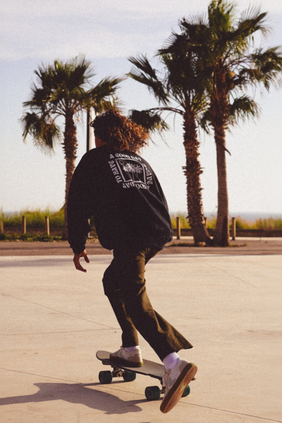 Mann fährt Skateboard am Meer und trägt einen schwarzen Oversized Sweater.