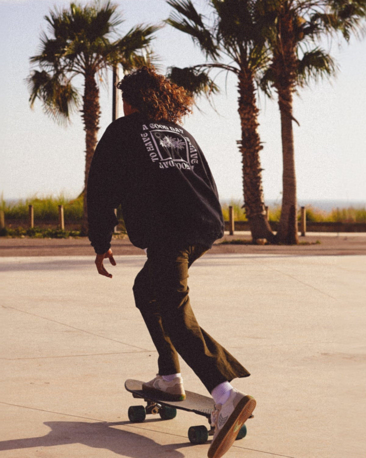 Mann fährt Skateboard am Meer und trägt einen schwarzen Oversized Sweater.