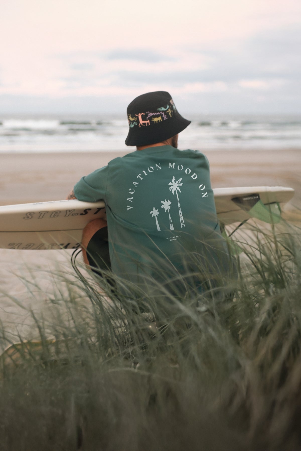 Mann sitzt am Strand in den Dünen, schaut auf das Meer und trägt das Vacation Mood On T-Shirt in Green Bay von The Sunkissed Rebels