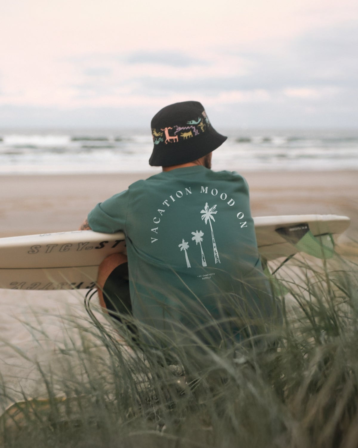 Mann sitzt am Strand in den Dünen, schaut auf das Meer und trägt das Vacation Mood On T-Shirt in Green Bay von The Sunkissed Rebels