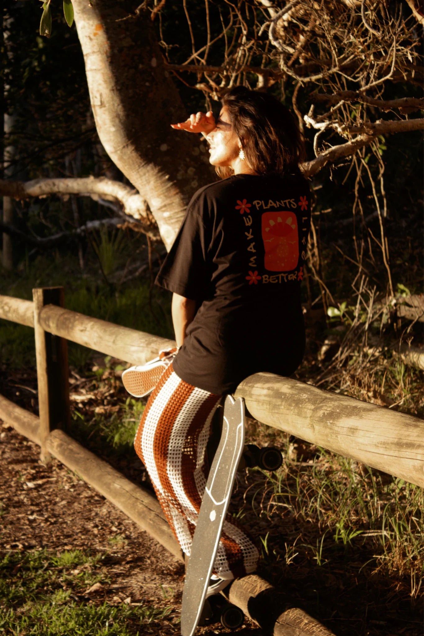 Frau sitzt auf Holzgeländer mit Skateboard und trägt schwarzes Better Life Light Oversized T-Shirt mit Rückenprint im Sonnenlicht