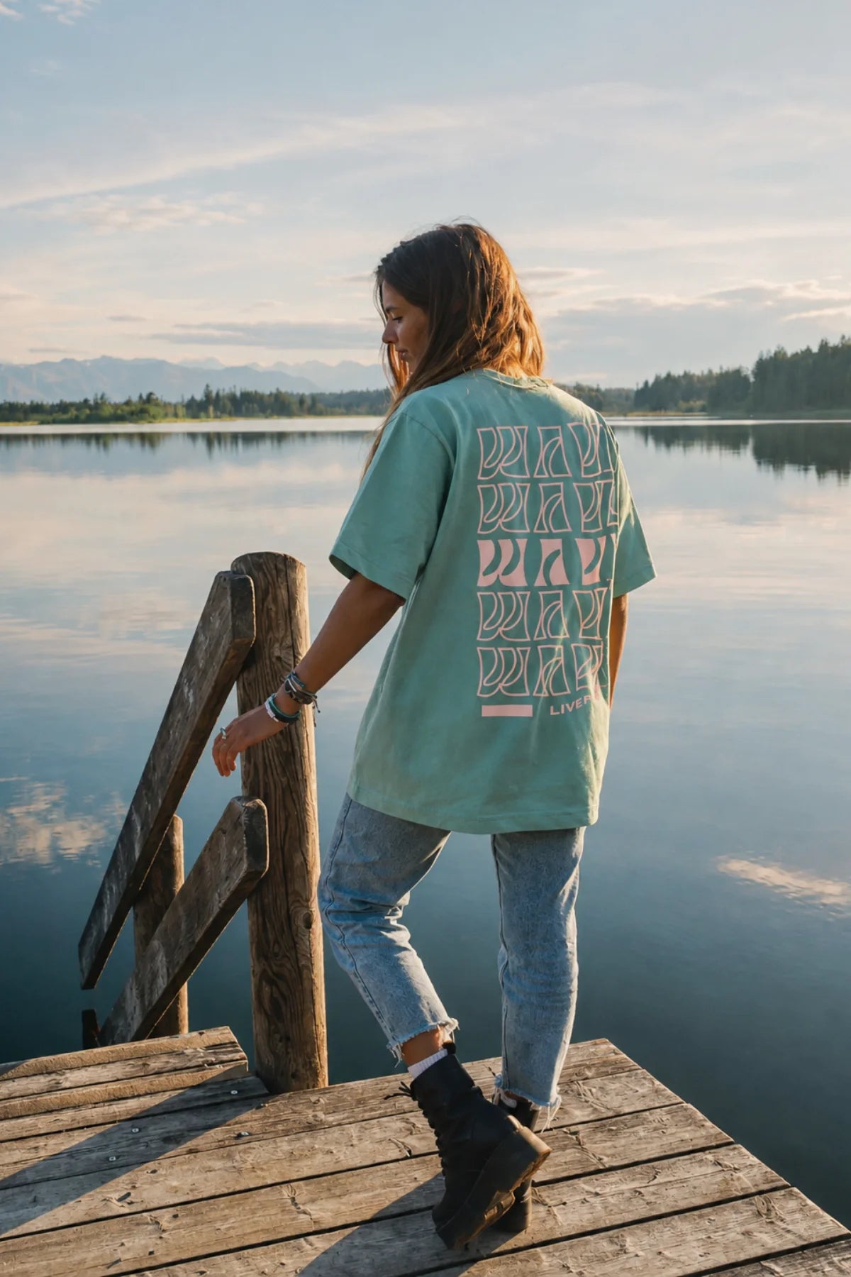 Rückenansicht des hellgrünen Live Free Oversized Shirts mit typografischem Wave-Print in Rosa auf einem Steg am Wasser.