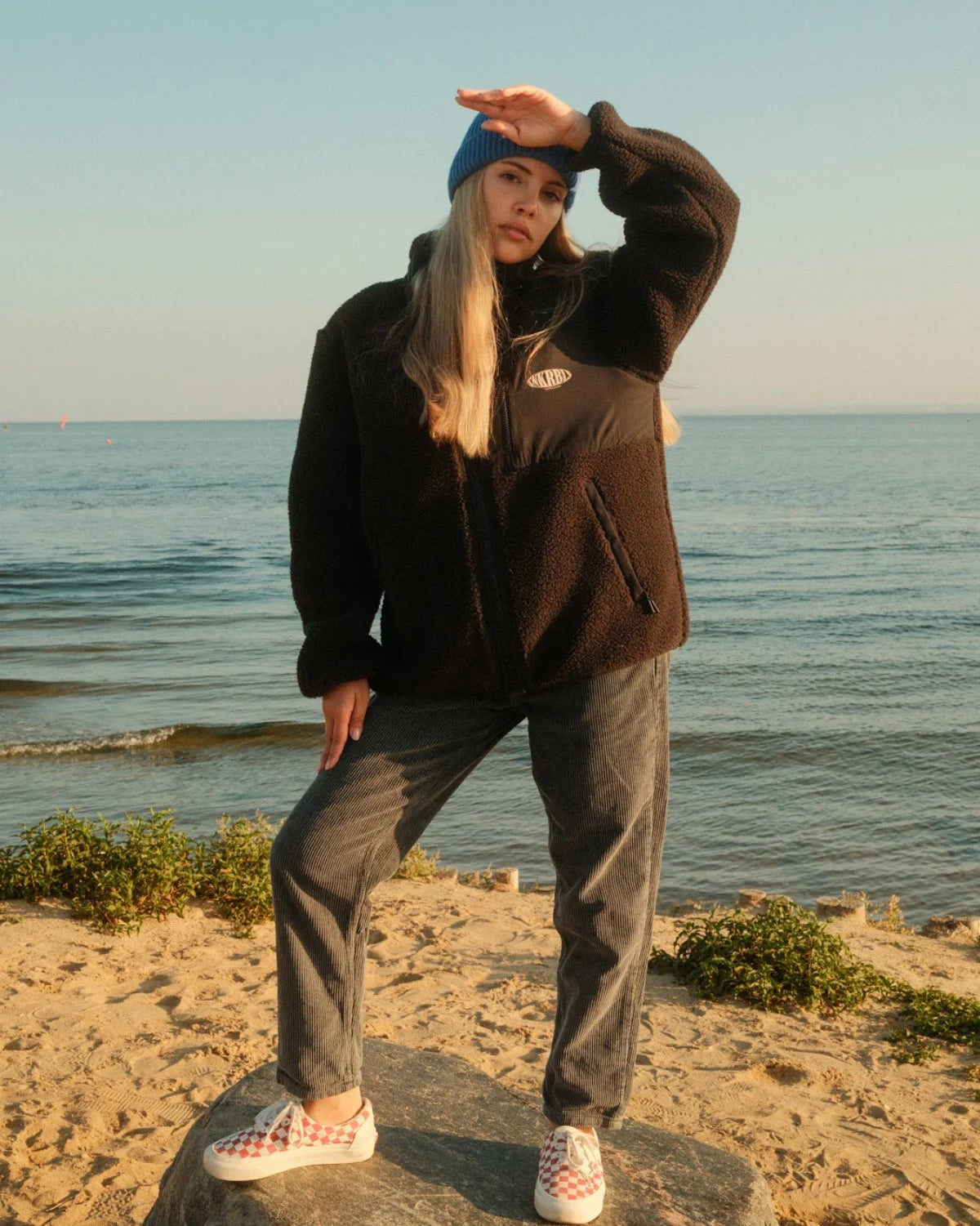 Schwarze Teddy Fleecejacke von The Sunkissed Rebels kombiniert mit Kordhose und blauer Beanie am Meer.