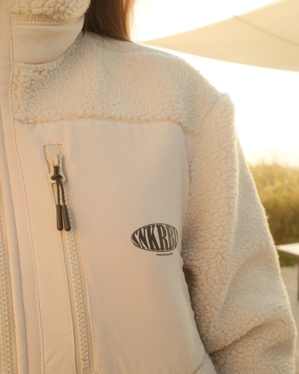 Detail der beigefarbenen Fleece Jacket von The Sunkissed Rebels mit Reißverschluss-Brusttasche und SNKRBL Logo-Print.