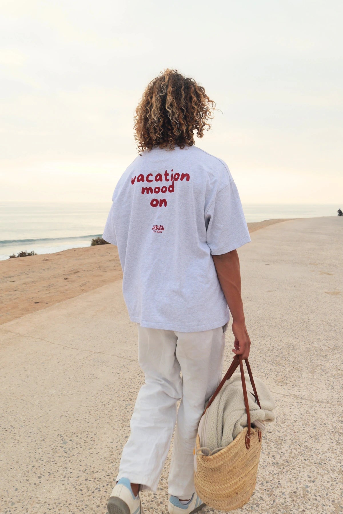 Person trägt das Goodlife Heavy T-Shirt in Cool Grey mit Vacation Mood On Rückenprint auf einer Küstenpromenade am Meer.