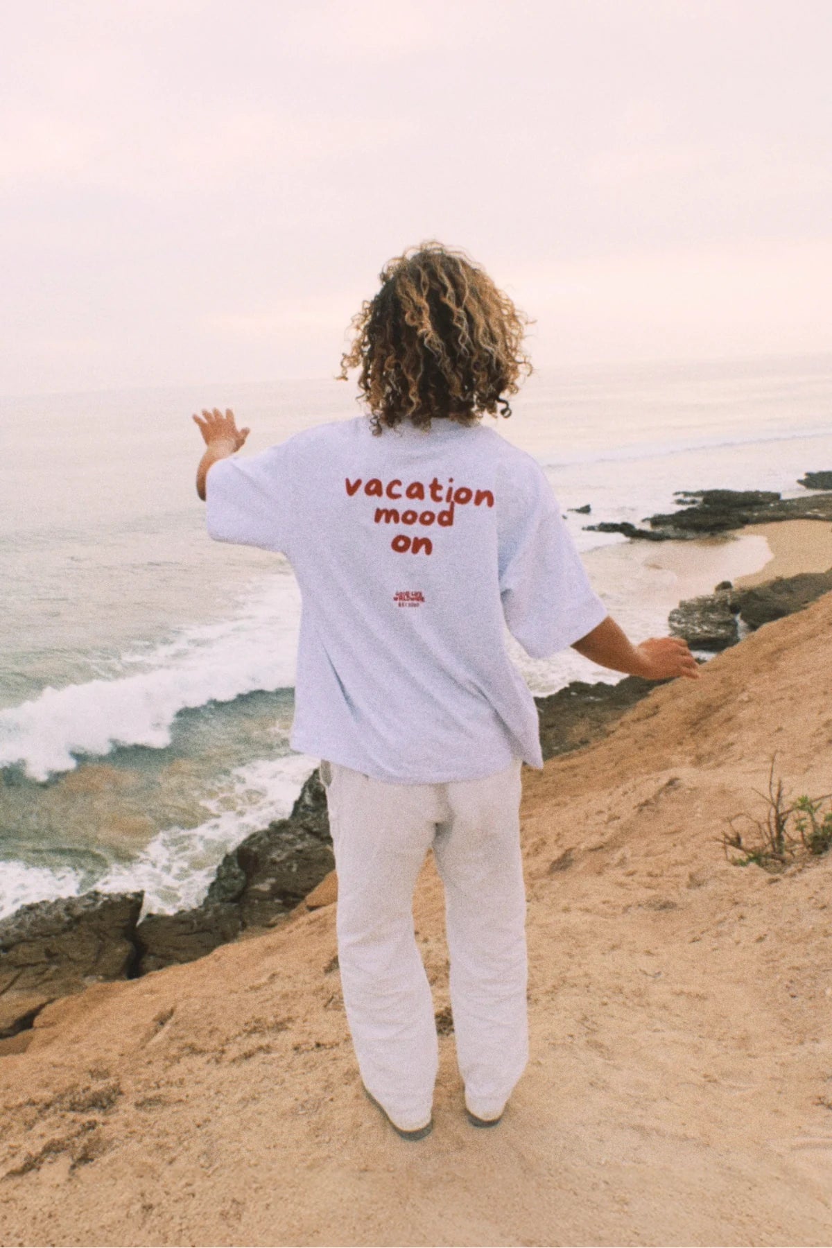 Person trägt das Goodlife Heavy T-Shirt in Cool Grey mit Vacation Mood On Print an einer Küstenklippe über dem Meer.