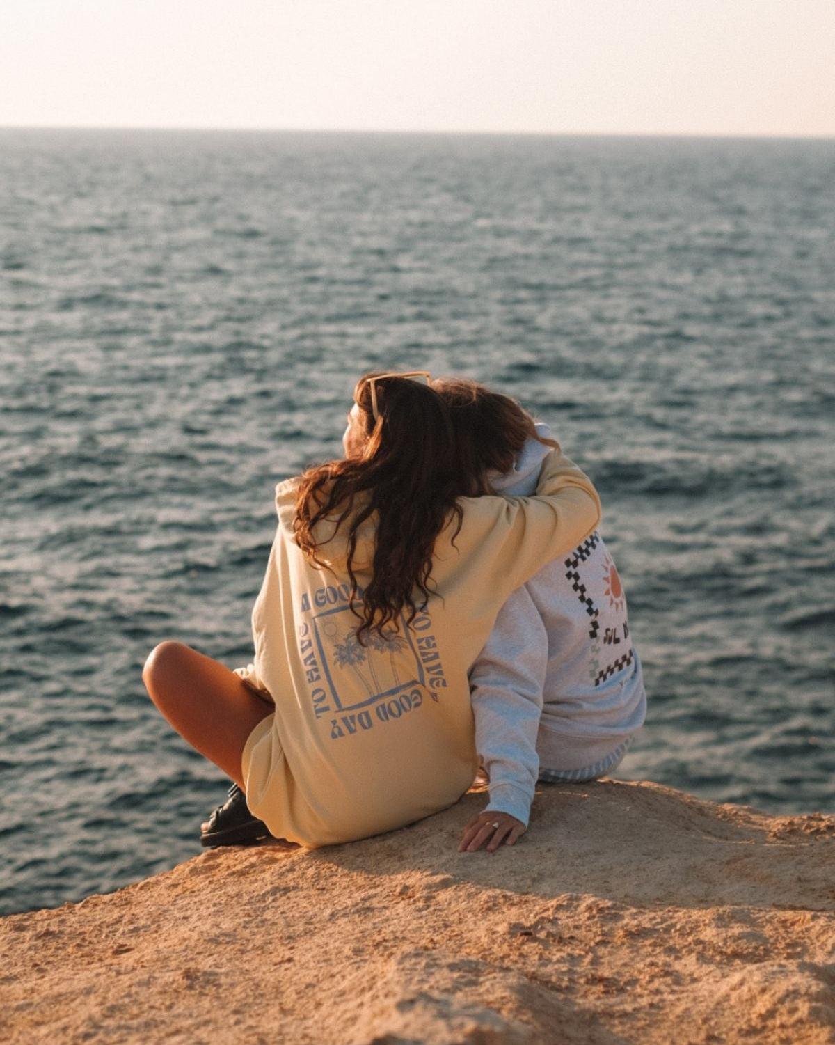 Zwei Frauen sitzen am Strand, halten sich in den Armen und schauen den Sonnenuntergang. Beide tragen Hoodies von The Sunkissed Rebels.