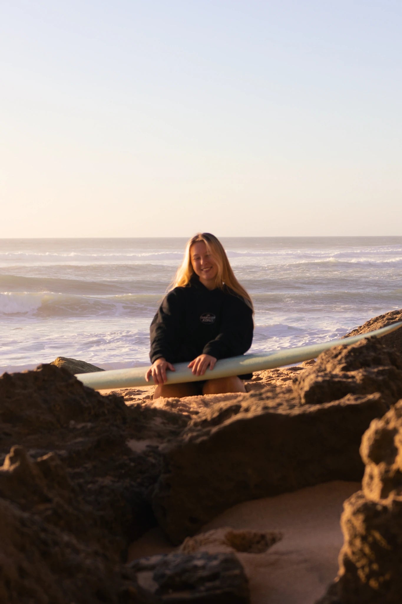 Frau sitzt am Strand mit Surfboard und trägt schwarzen Oversized Hoodie bei warmem Sonnenuntergang am Meer