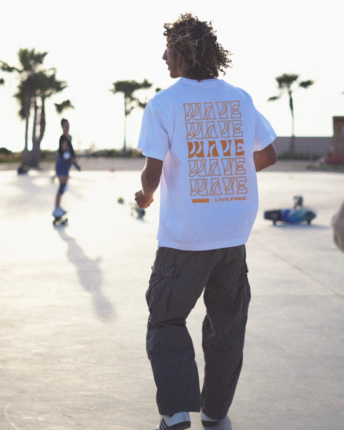 Rückansicht eines Skaters im weißen Oversized T-Shirt mit orangem Live Free Design – The Sunkissed Rebels