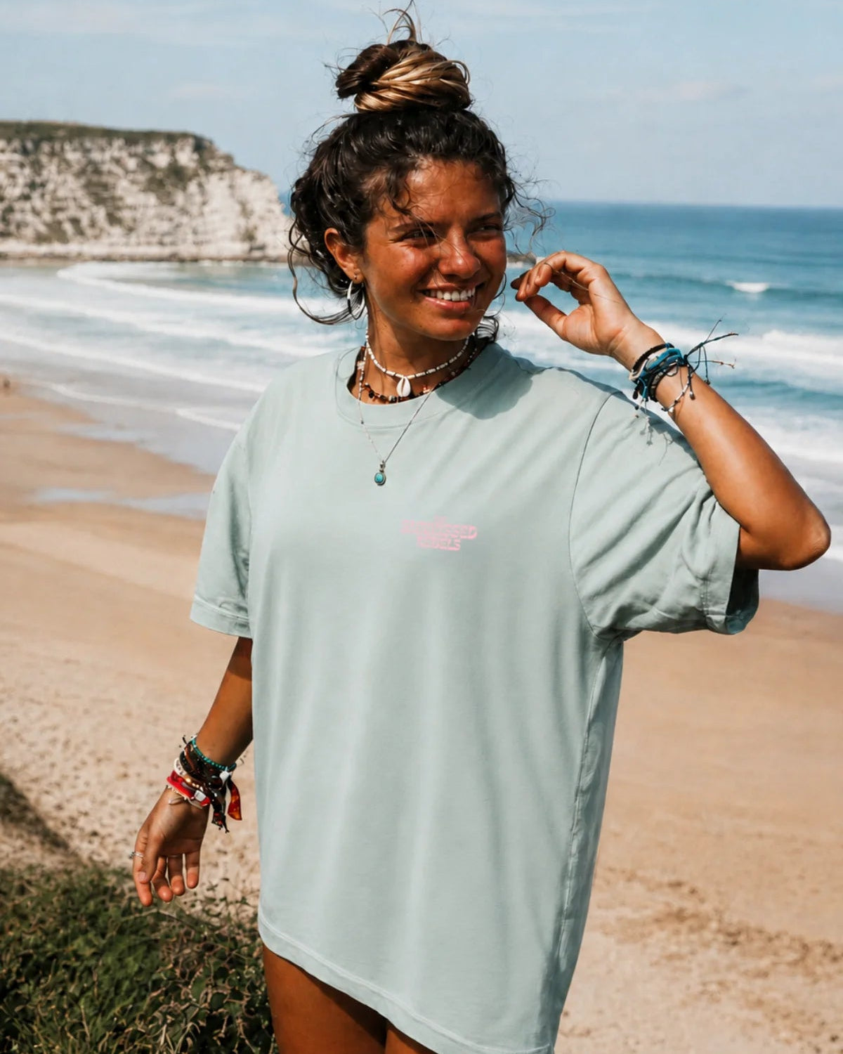 Lächelnde junge Frau am Strand im mintgrünen Live Free Oversized T-Shirt mit kleinem Brustprint.