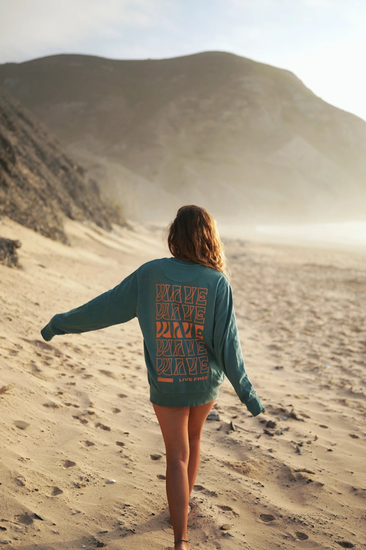 Frau läuft mit ausgestrecktem Arm im Palm Green Sweater am Meer entlang