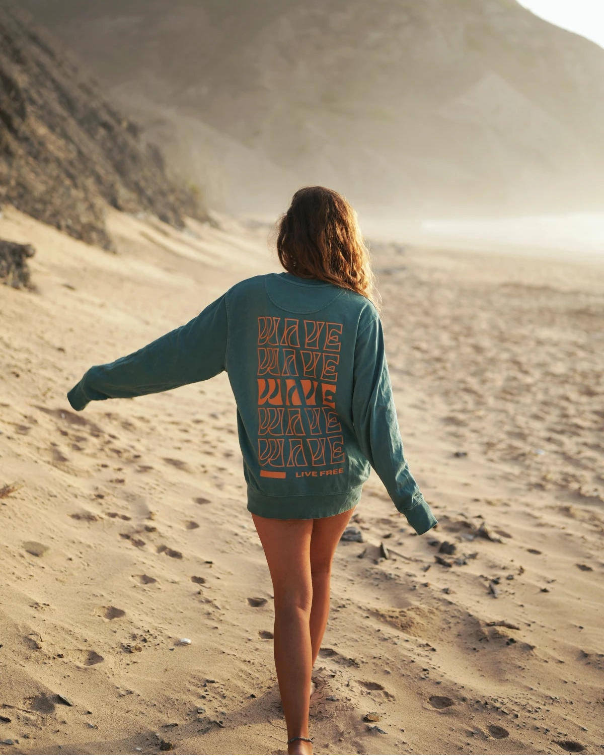 Frau läuft mit ausgestrecktem Arm im Palm Green Sweater am Meer entlang