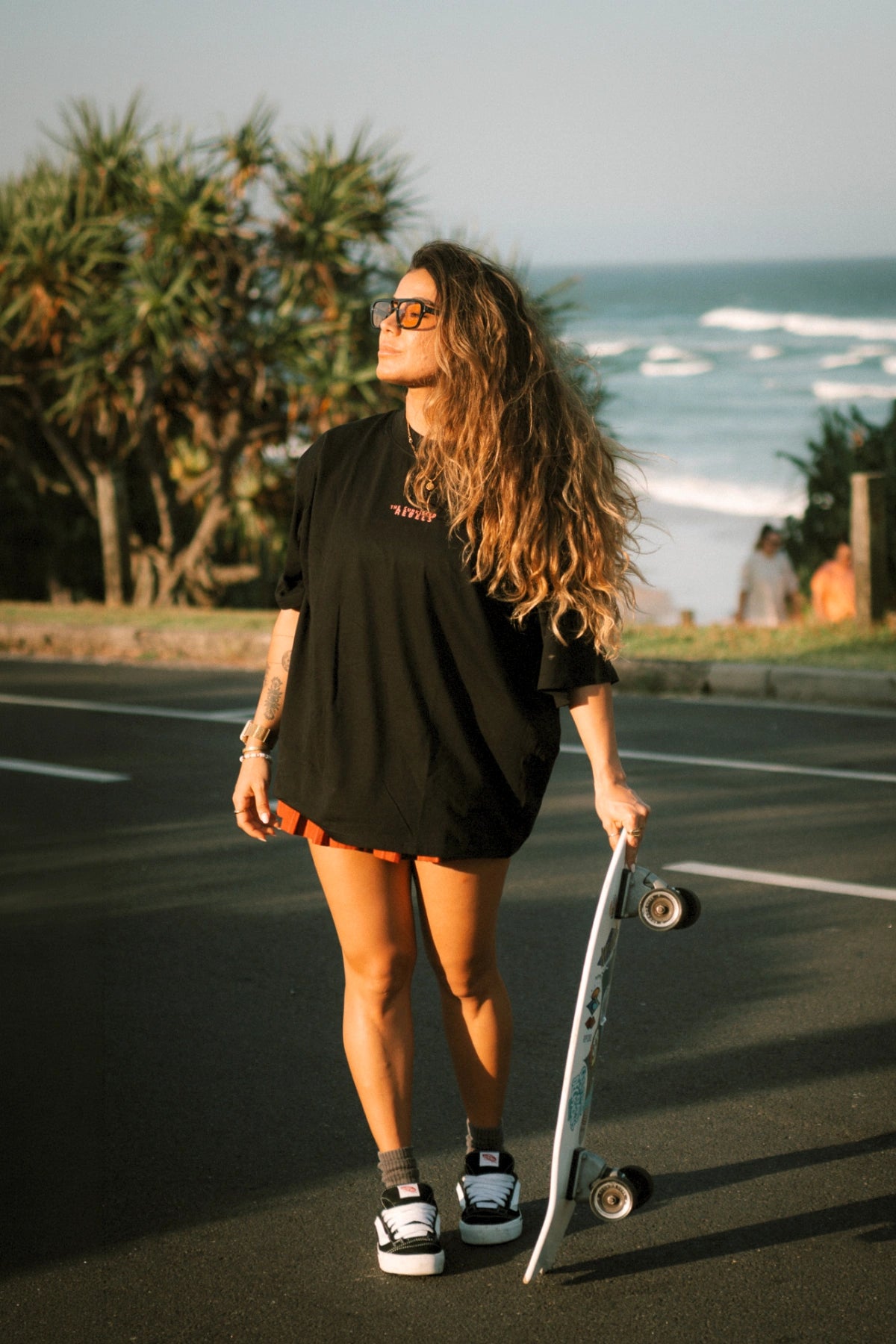 Frau im schwarzen No Bullshit Whale Boxy Oversized T-Shirt von The Sunkissed Rebels mit Skateboard an der Küste.