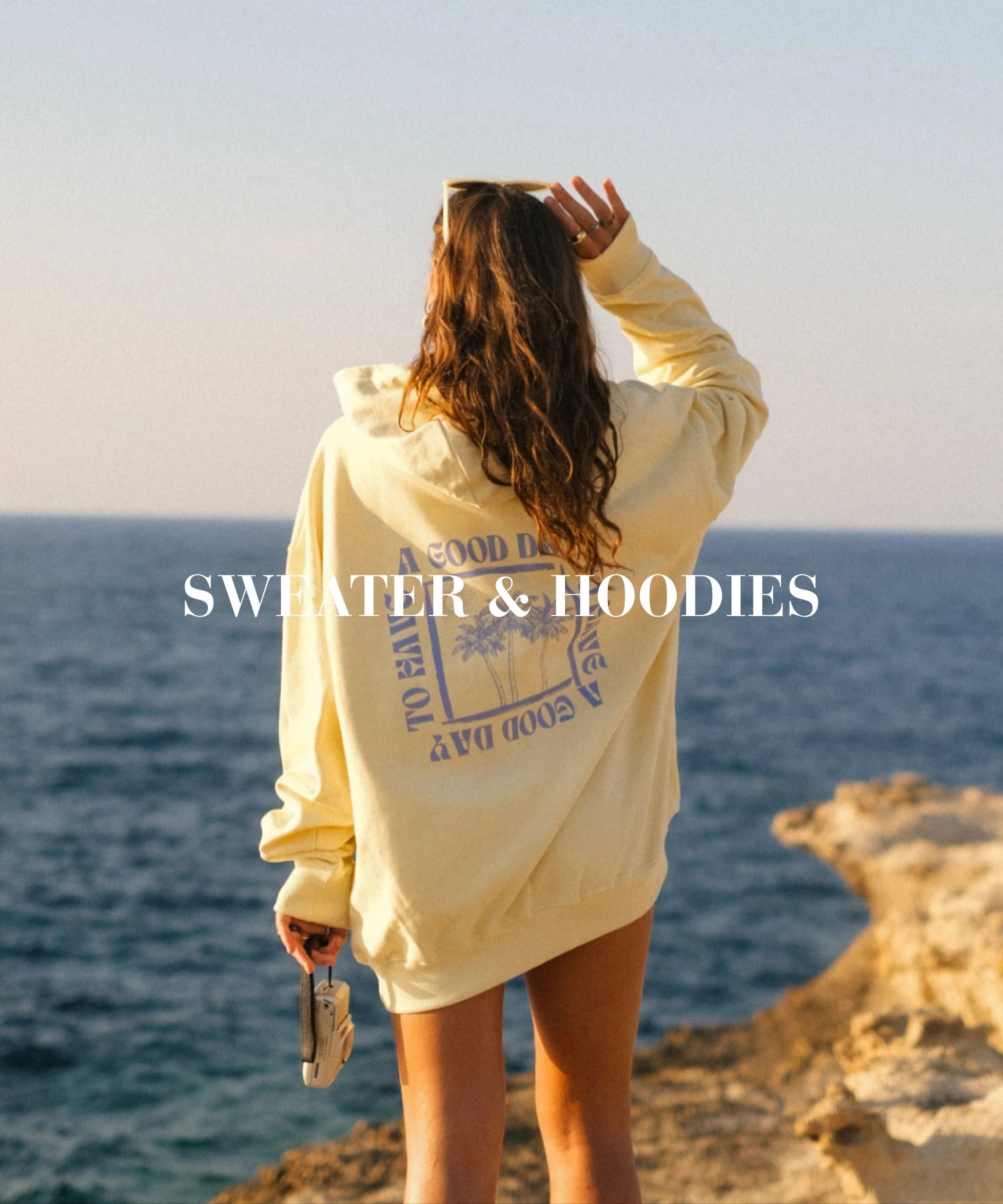 Frau steht am Felsen mit Blick aufs Meer und trägt einen gelben Oversized Hoodie mit Palmen-Print – Sweater & Hoodies Kollektion von The Sunkissed Rebels