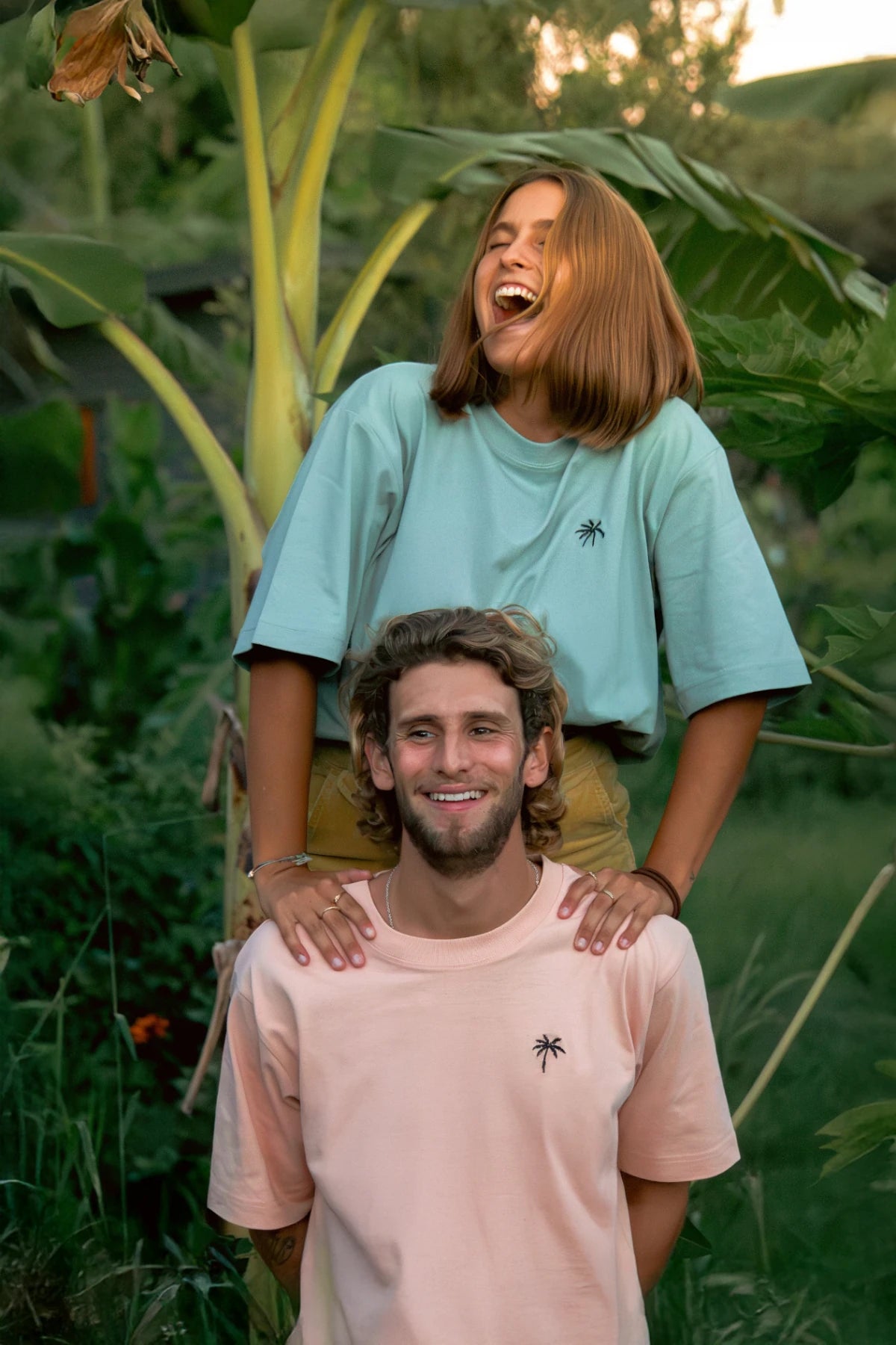 Frau und Mann tragen fröhlich Oversized T-Shirts mit Palmenstick unter Bananenstaude