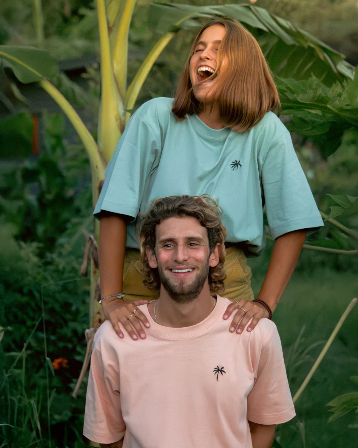Frau und Mann tragen fröhlich Oversized T-Shirts mit Palmenstick unter Bananenstaude