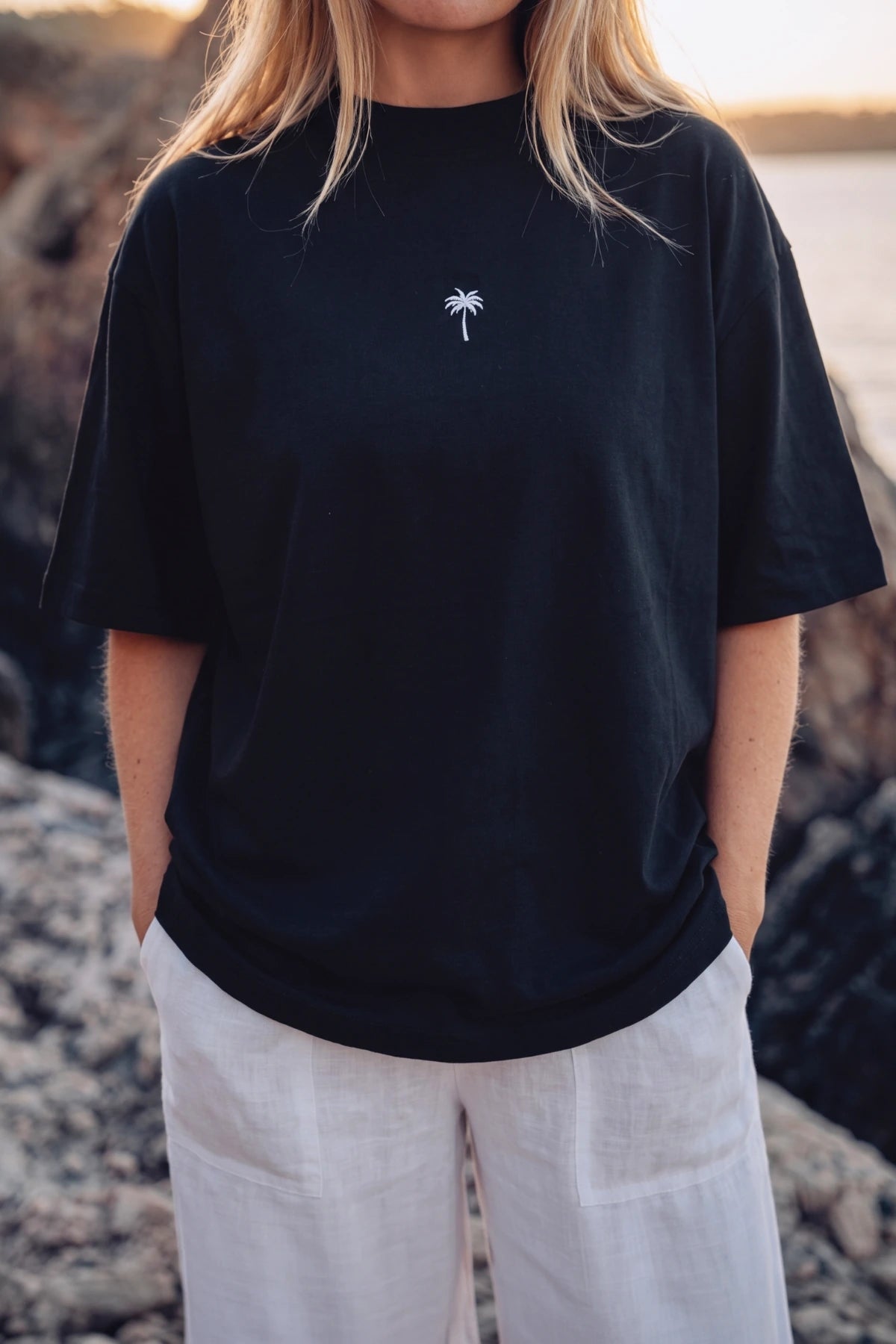 Frau trägt das Palm Tree Boxy Oversized T-Shirt Black mit lockerer Passform an einer Küste mit Felsen und Meer im Hintergrund.