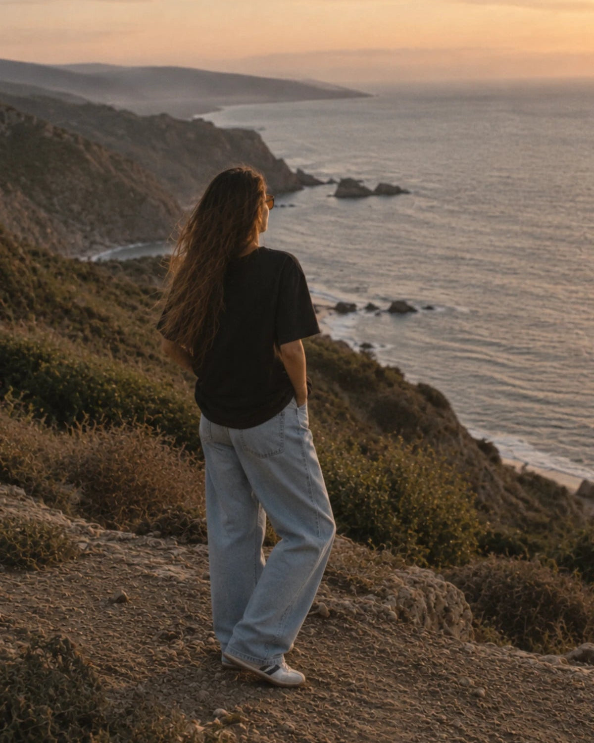Rückenansicht des schwarzen Oversized Shirts von The Sunkissed Rebels im warmen Sonnenuntergang an den Klippen.