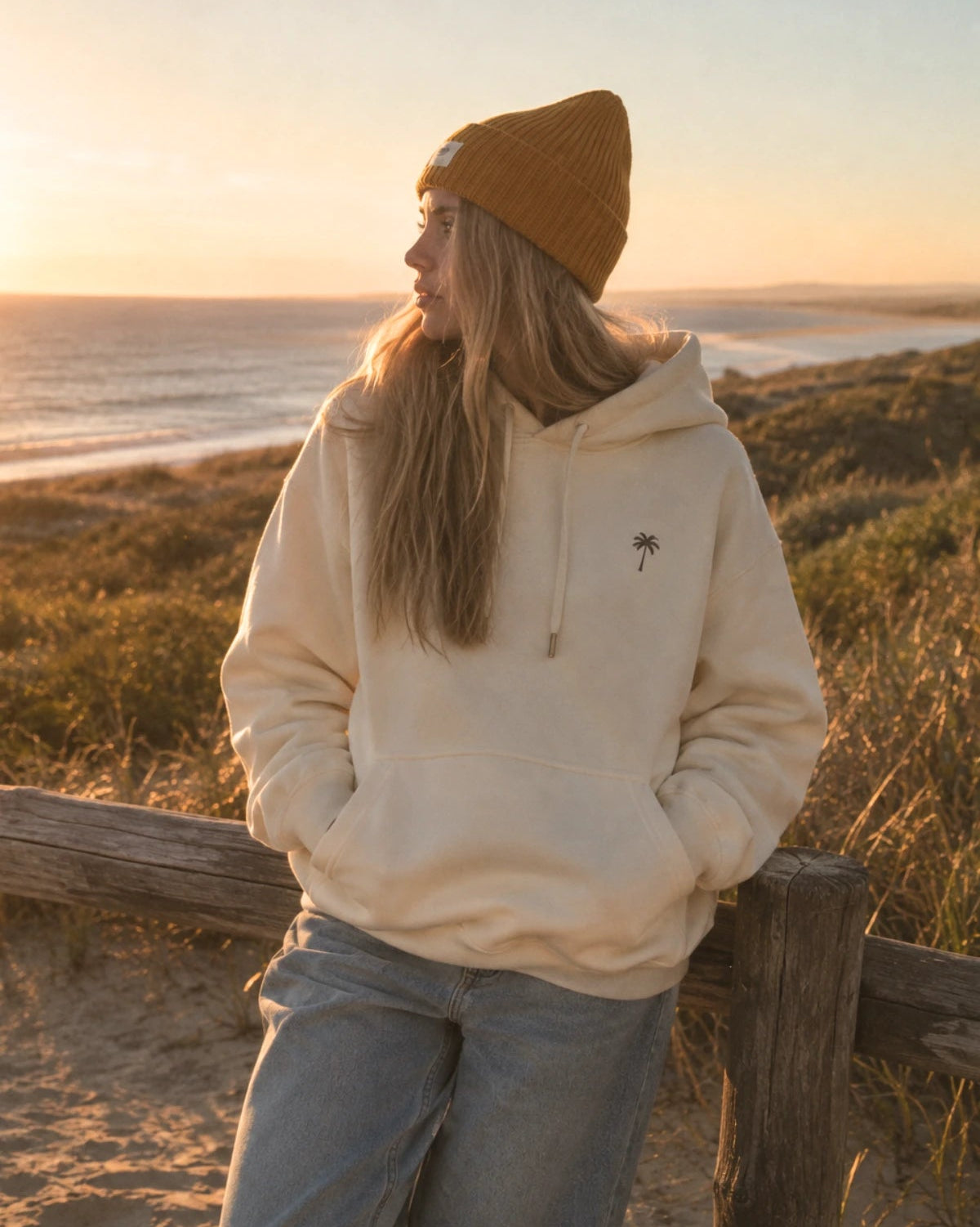 Frau am Strand mit cremefarbenem Kapuzenpullover Palm Tree Hoodie in Natural Raw von The Sunkissed Rebels, Kängurutasche und gelber Beanie.