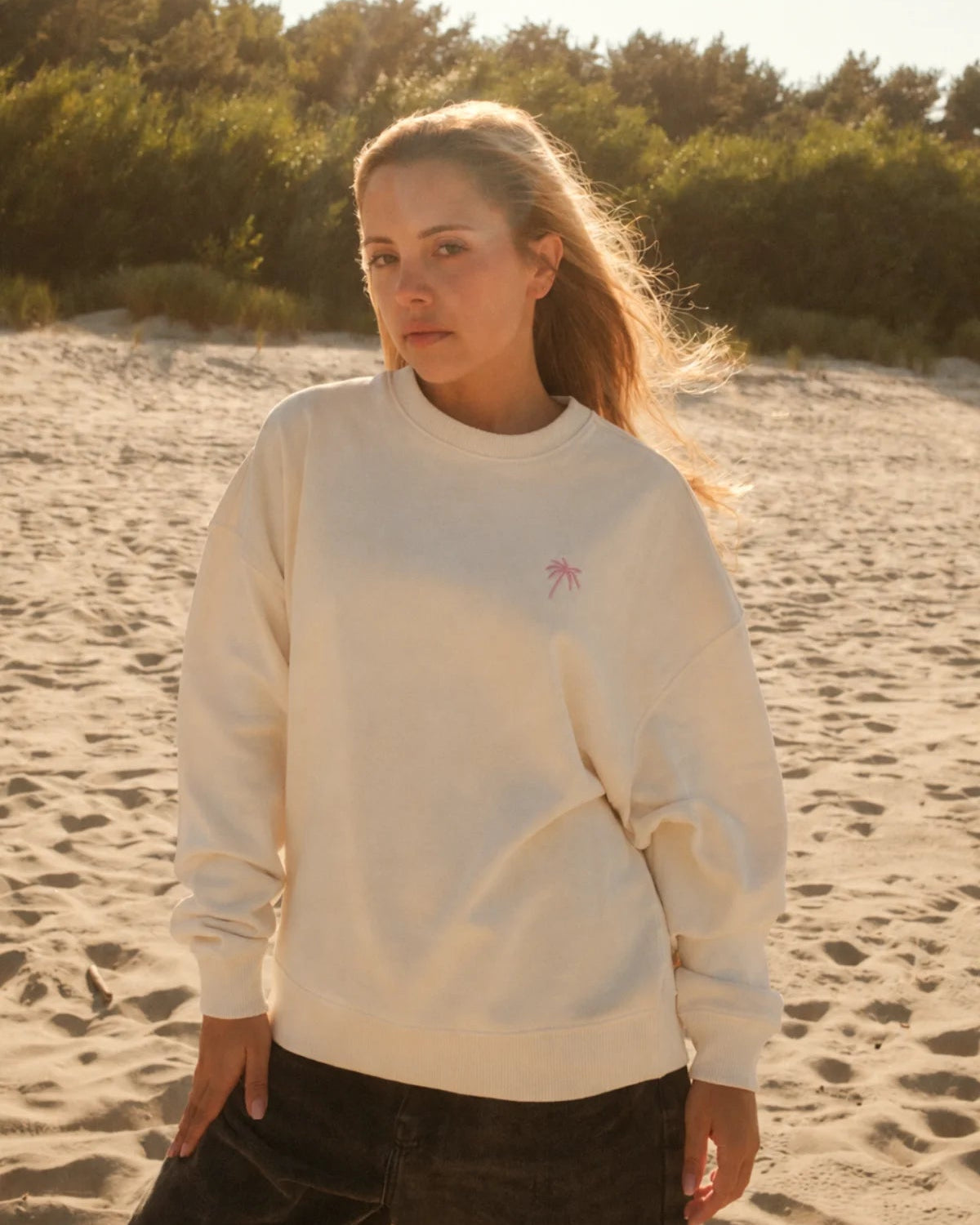 Frau am Strand im beigen Palm Tree Oversized Sweater in Cream von The Sunkissed Rebels mit rosa Palmen-Stickerei.