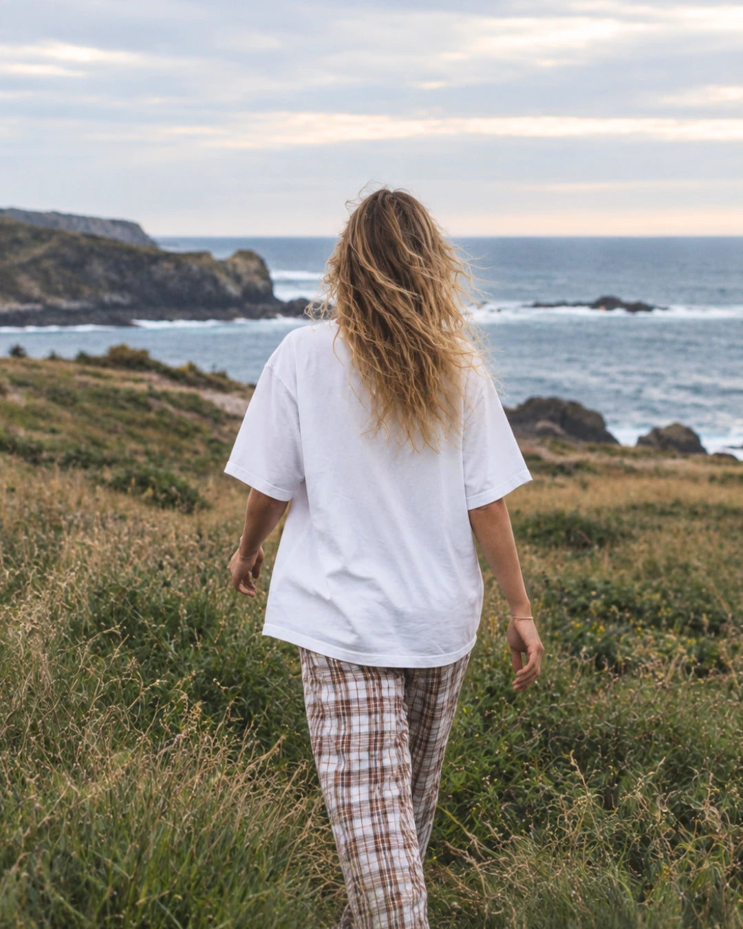 Rückansicht einer Frau im weißen Palm Tree Oversized T-Shirt auf einer Küstenwiese mit Blick auf das Meer.