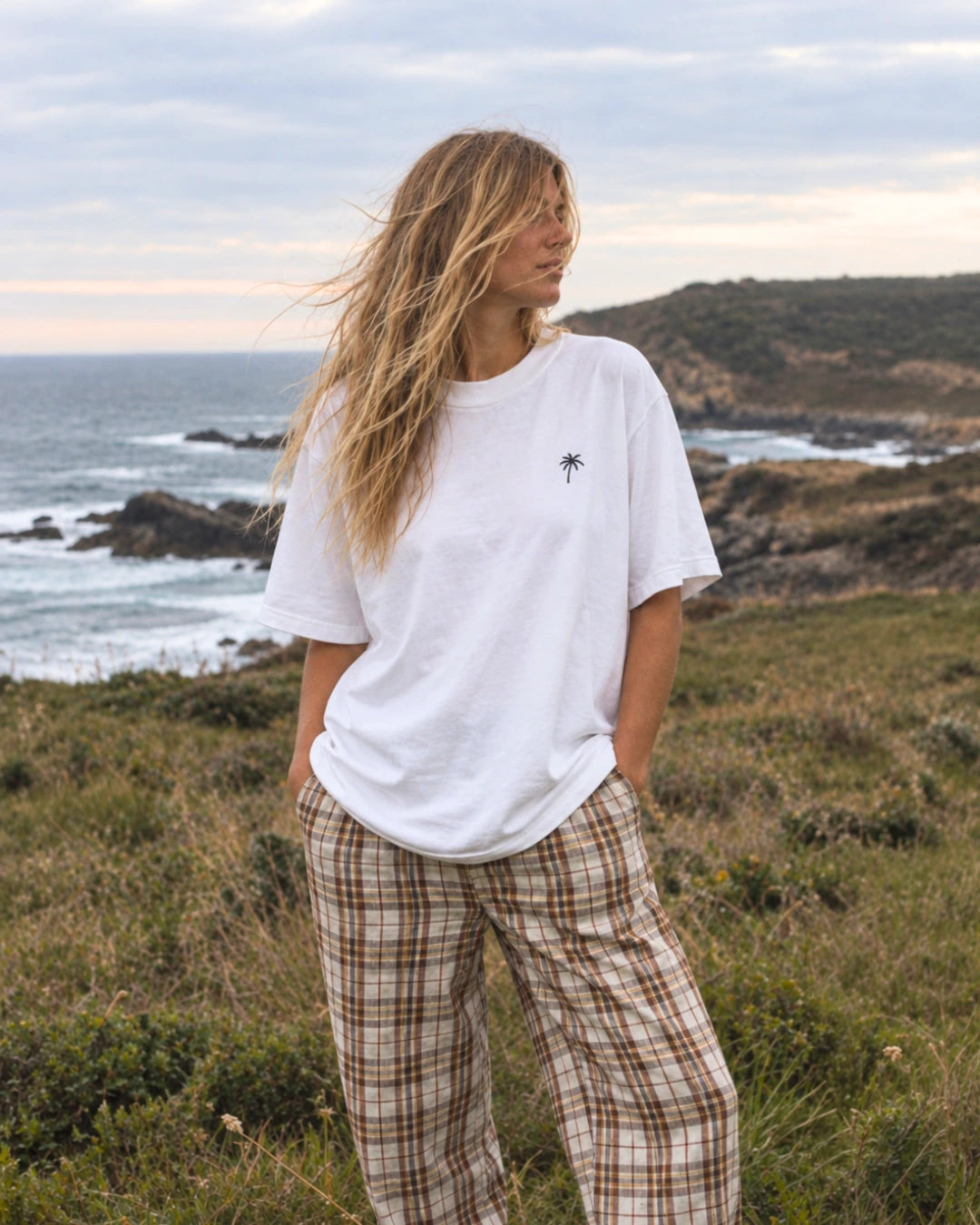 Frau trägt das weiße Palm Tree Oversized T-Shirt von The Sunkissed Rebels an einer felsigen Küste mit Meer im Hintergrund.