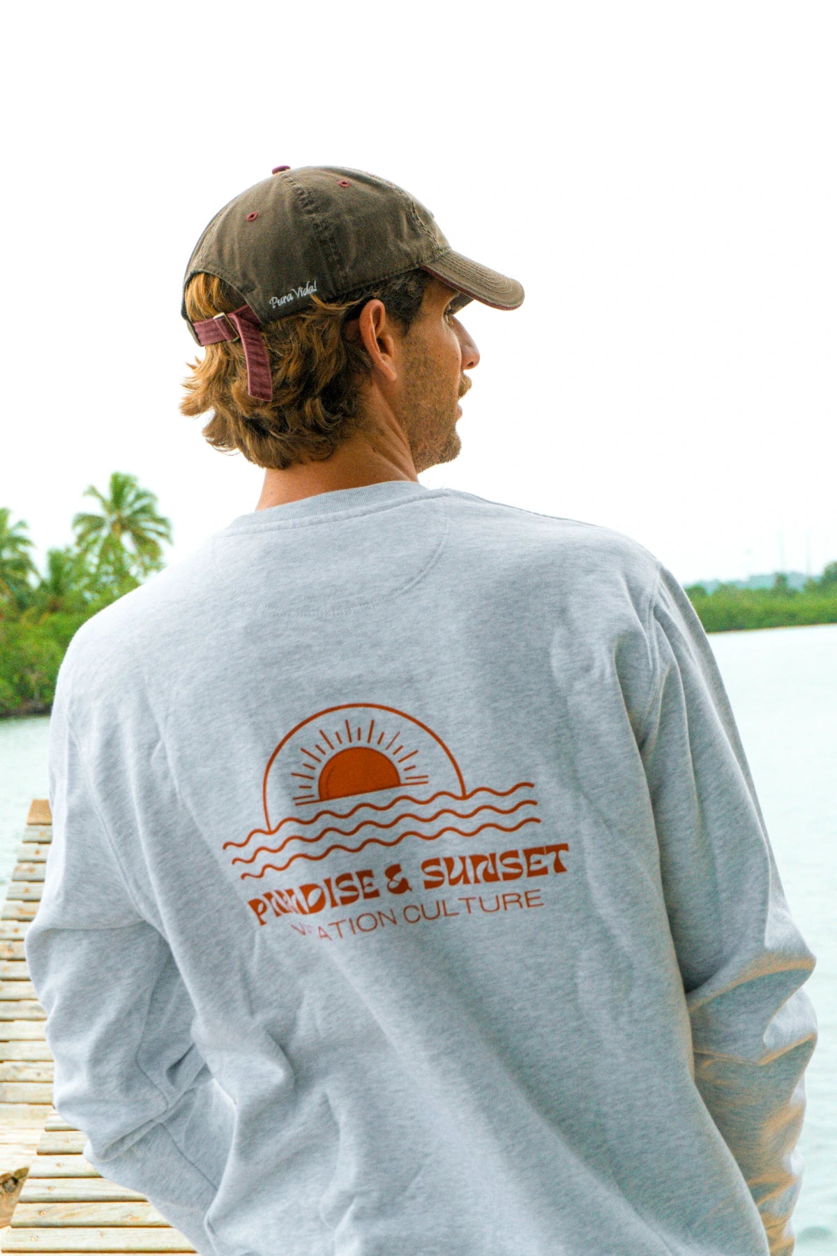 Schräg aufgenommene Rückansicht des Paradise & Sunset Sweaters in Cool Grey mit großem Paradise & Sunset Print am Wasser.