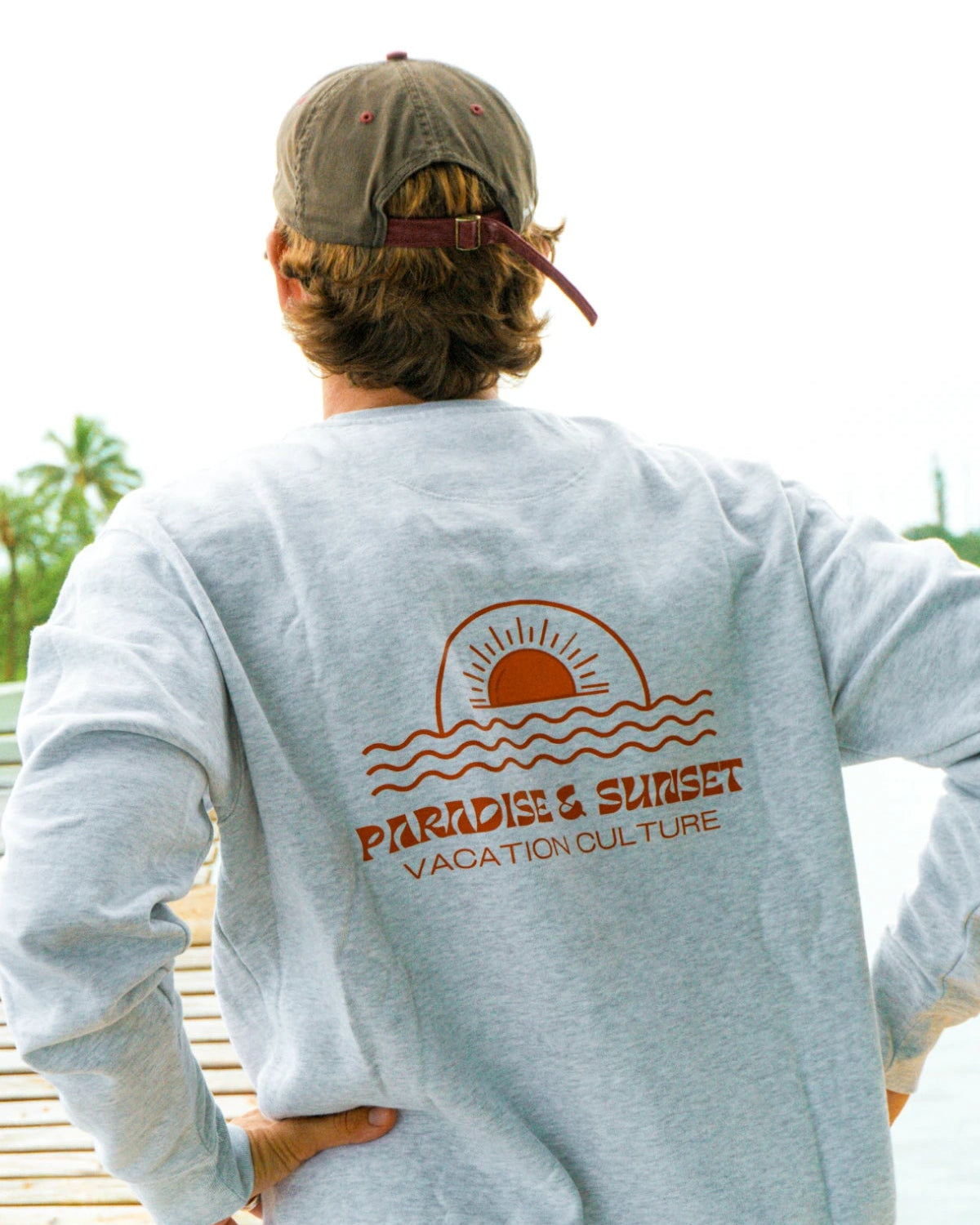 Rückansicht des Paradise & Sunset Sweaters in Cool Grey mit großem Paradise & Sunset Rückenprint auf einem Steg am tropischen Wasser.