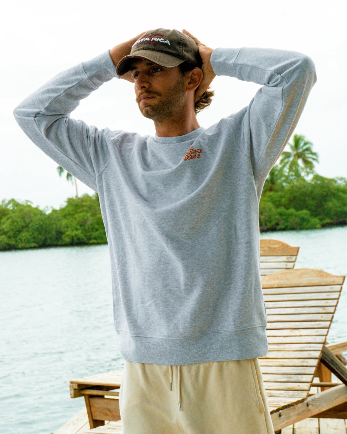 Mann trägt den Paradise & Sunset Sweater in Cool Grey auf einem Holzsteg am tropischen Wasser mit entspannter Vacation-Vibes-Atmosphäre.