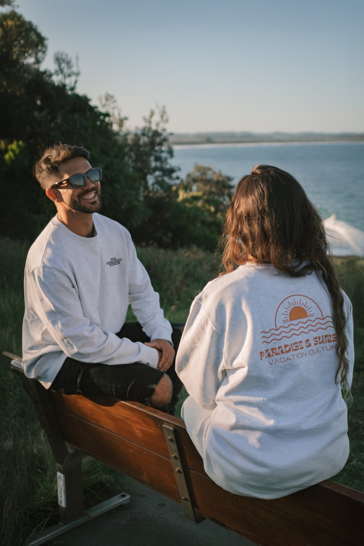 Mann und Frau sitzen auf einer Bank am Meer in Australien und tragen die Sweater der Paradise & Sunset Kollektion von The Sunkissed Rebels.