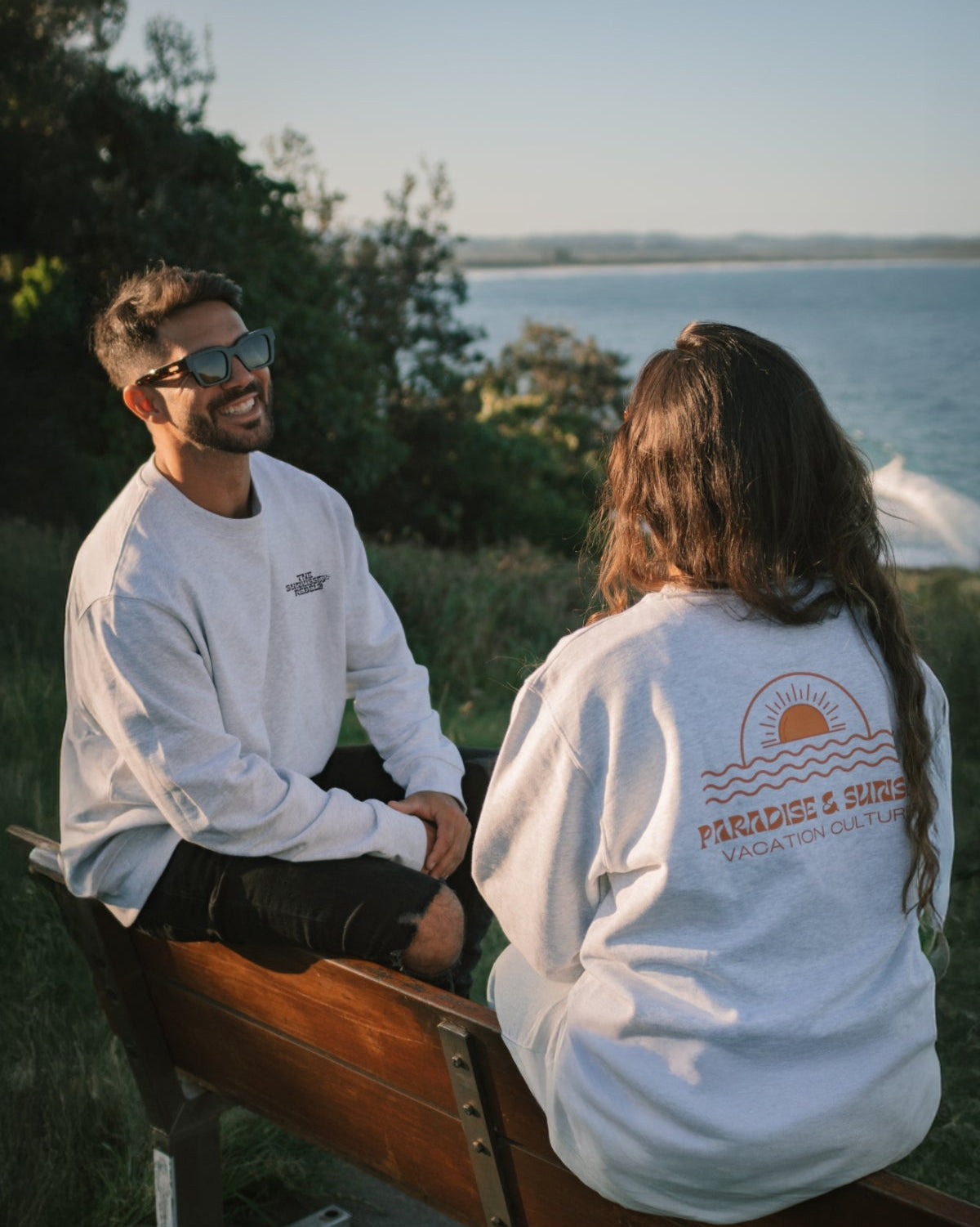 Mann und Frau sitzen auf einer Bank am Meer in Australien und tragen die Sweater der Paradise & Sunset Kollektion von The Sunkissed Rebels.
