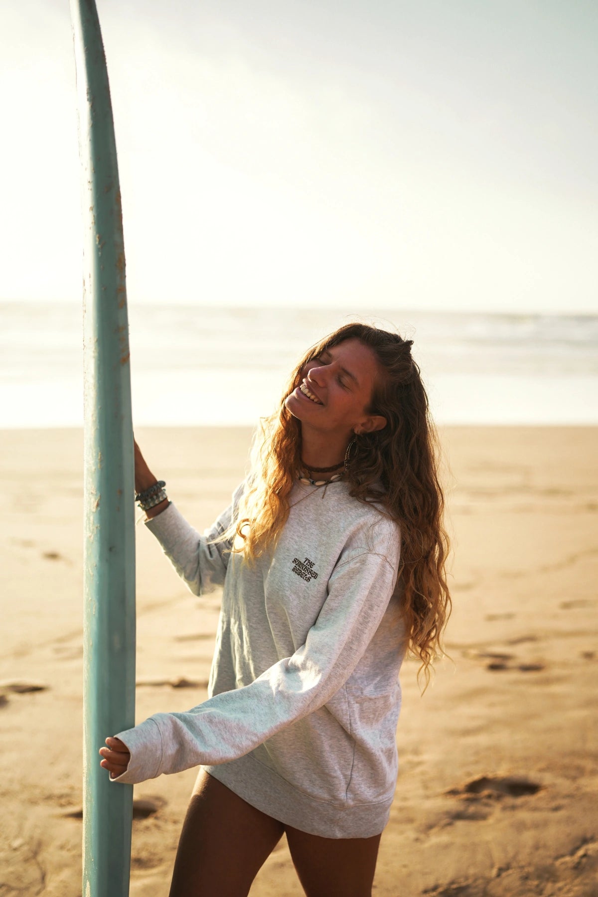 Frau mit Surfbrett lacht im grauen Paradise & Sunset Sweater am Meer
