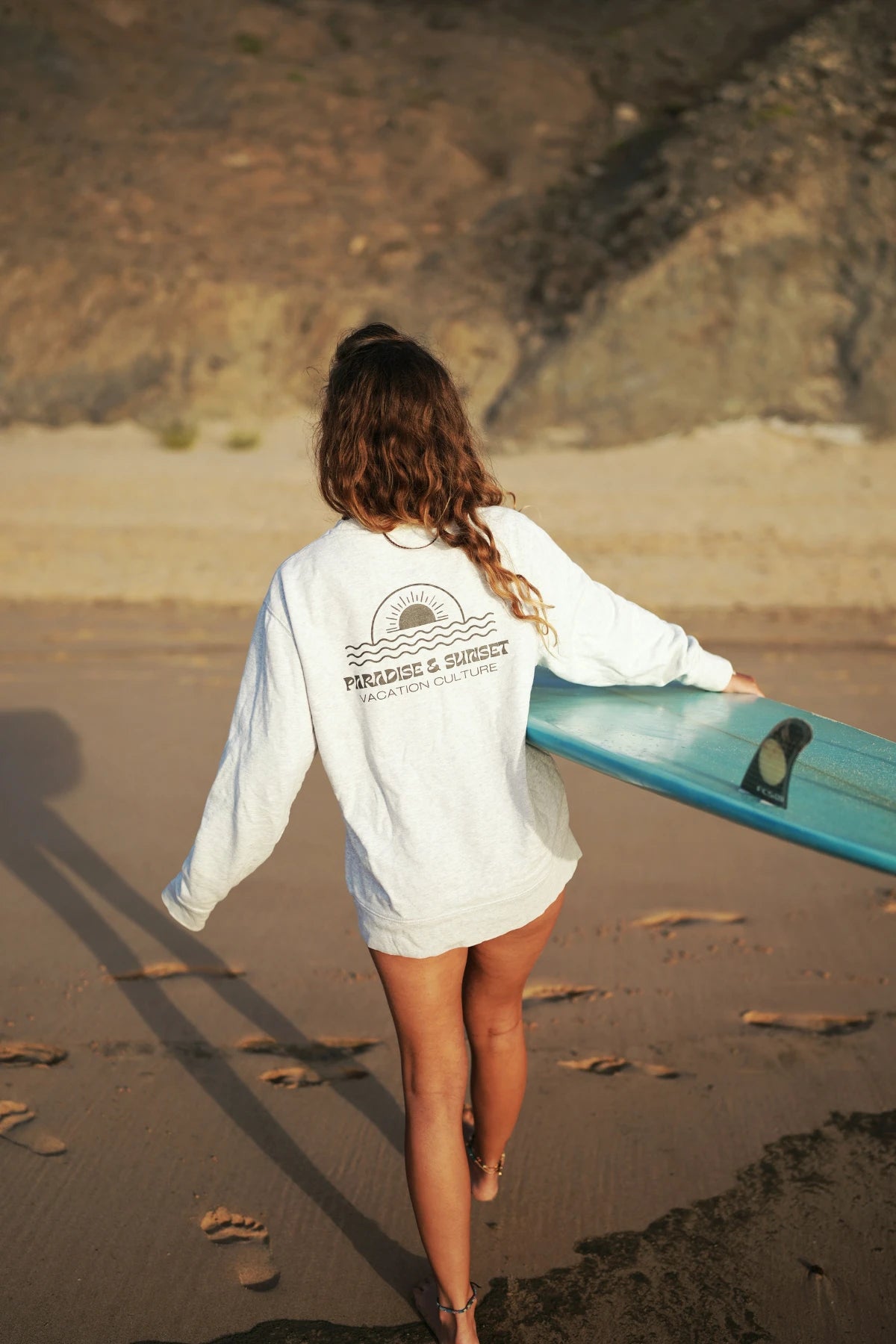 Frau mit Surfbrett in grauem Paradise & Sunset Sweater am Ozean