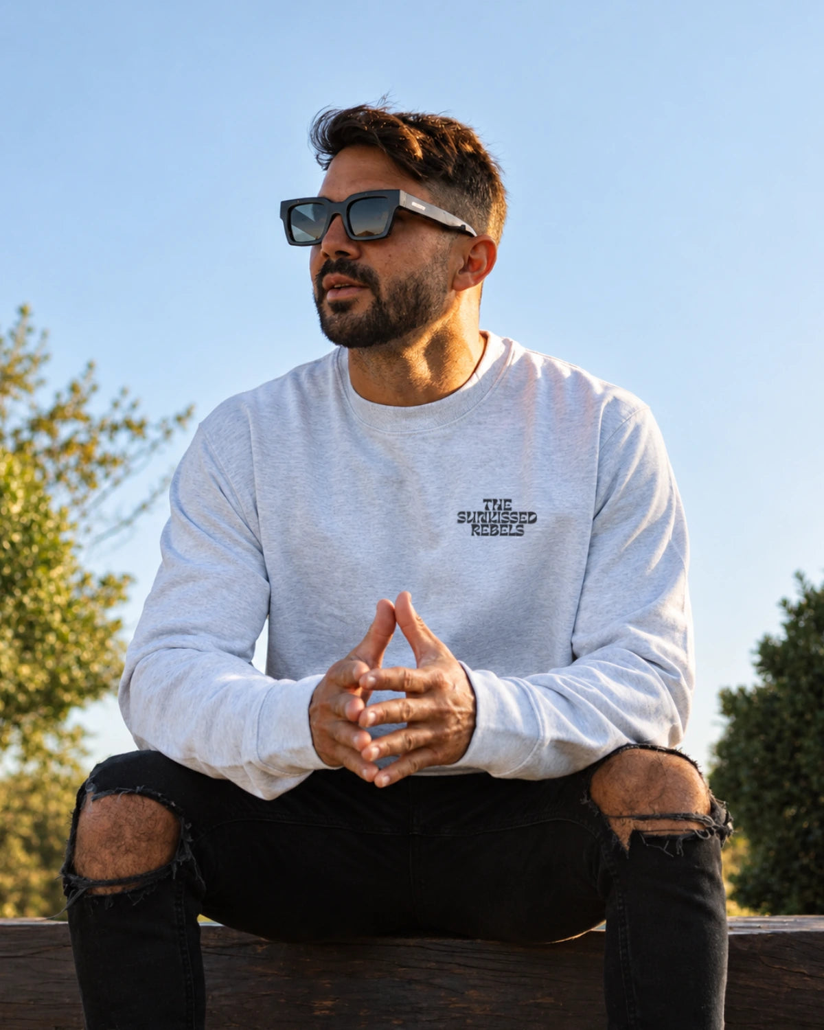 Mann trägt den Paradise & Sunset Sweater in Cool Grey mit kleinem The Sunkissed Rebels Brustprint