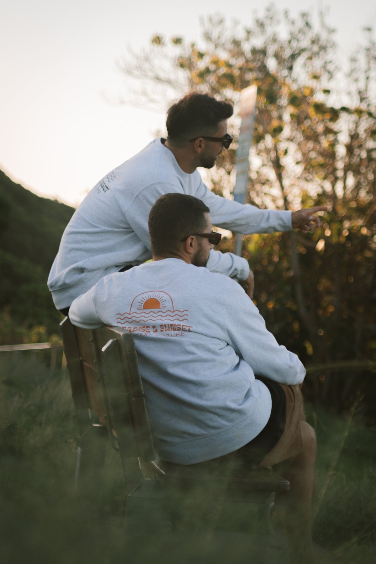 Zwei Männer sitzen auf einer Bank in Australien und tragen jeweils den Paradise & Sunset Sweater von The Sunkissed Rebels, einmal mit schwarzem und einmal mit orangenem Druck.