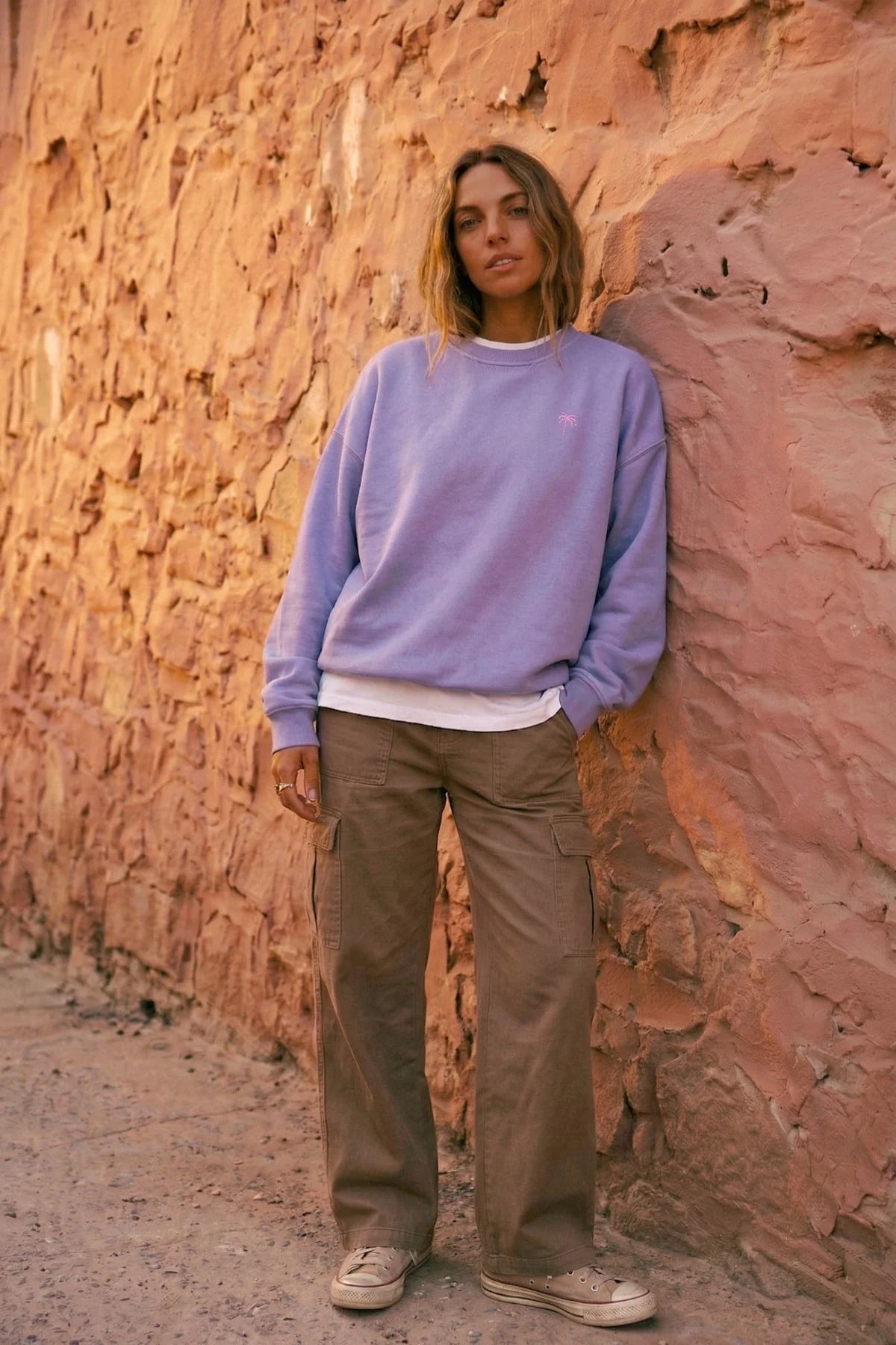 Damen in fliederfarbenem Oversized Vintage Sweater von The Sunkissed Rebels, lehnend an einer strukturierten Steinwand. Der Pullover hat eine kleine rosa Palmen-Stickerei.