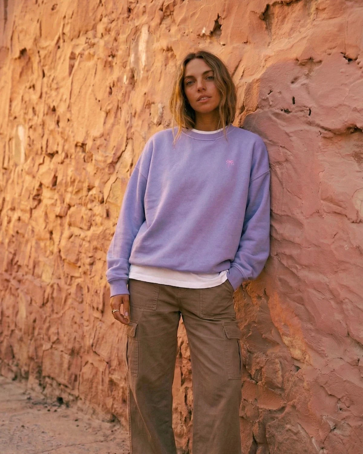 Damen in fliederfarbenem Oversized Vintage Sweater von The Sunkissed Rebels, lehnend an einer strukturierten Steinwand. Der Pullover hat eine kleine rosa Palmen-Stickerei.