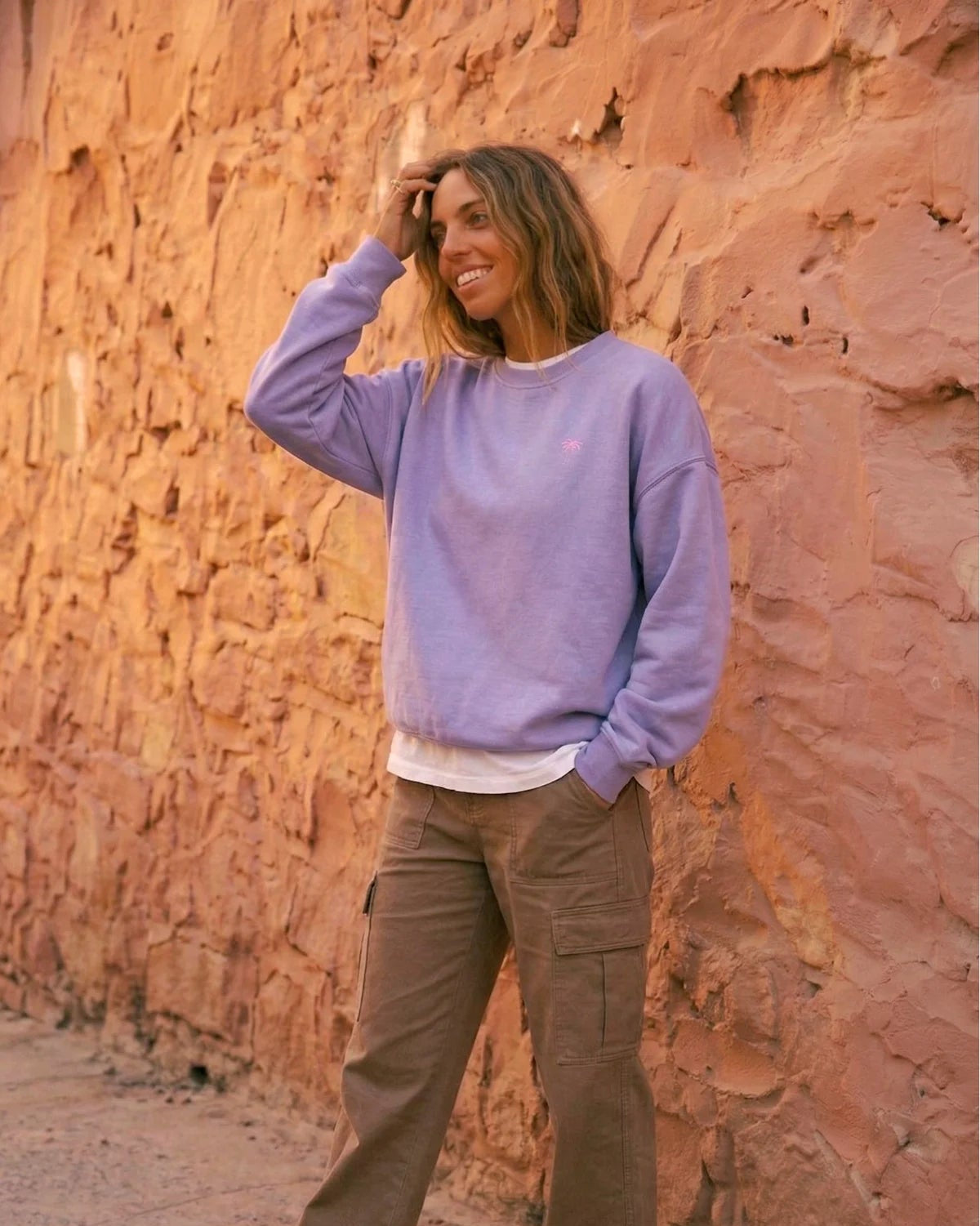 Frau lächelt und posiert im fliederfarbenen Pink Palm Oversized Vintage Sweater von The Sunkissed Rebels. Zeigt den lockeren Fit und das Palmen-Logo.