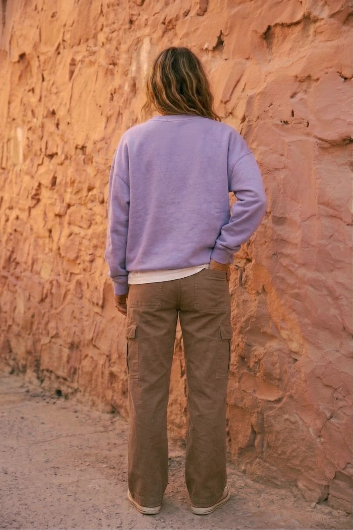 Rückansicht einer Frau im fliederfarbenen Oversized Vintage Sweater von The Sunkissed Rebels vor einer rustikalen Steinfassade. Betont den lockeren Sitz und die Farbe.