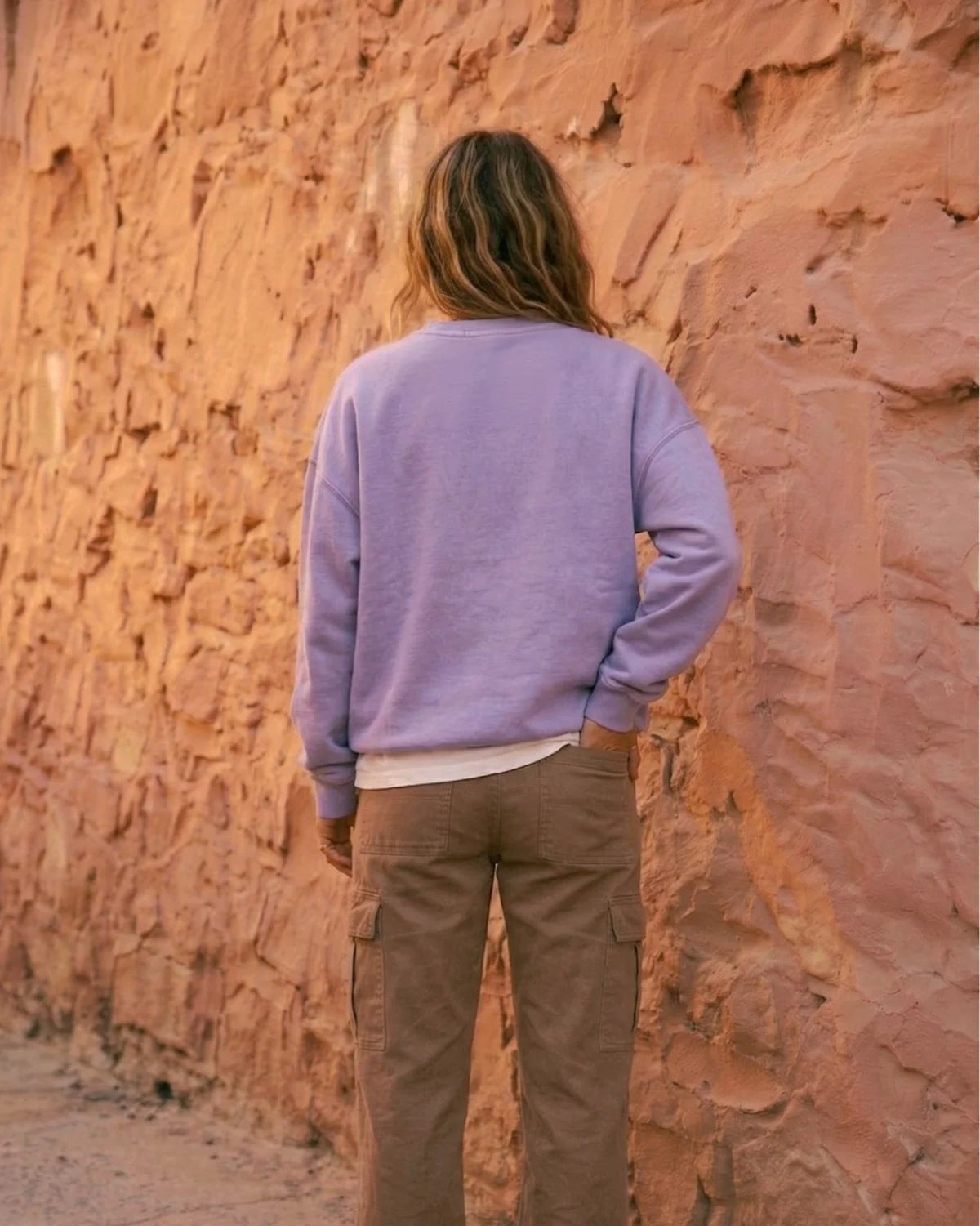 Rückansicht einer Frau im fliederfarbenen Oversized Vintage Sweater von The Sunkissed Rebels vor einer rustikalen Steinfassade. Betont den lockeren Sitz und die Farbe.