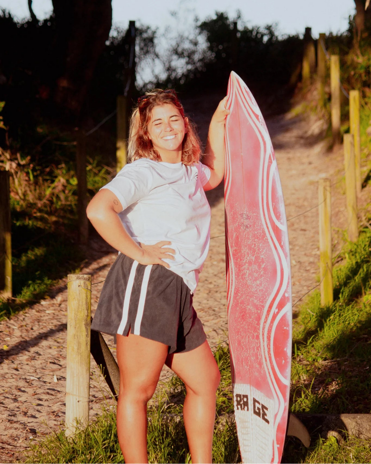 Frau mit Surfboard am Strandweg trägt weißes Rebel Elements Light T-Shirt im Sonnenuntergang