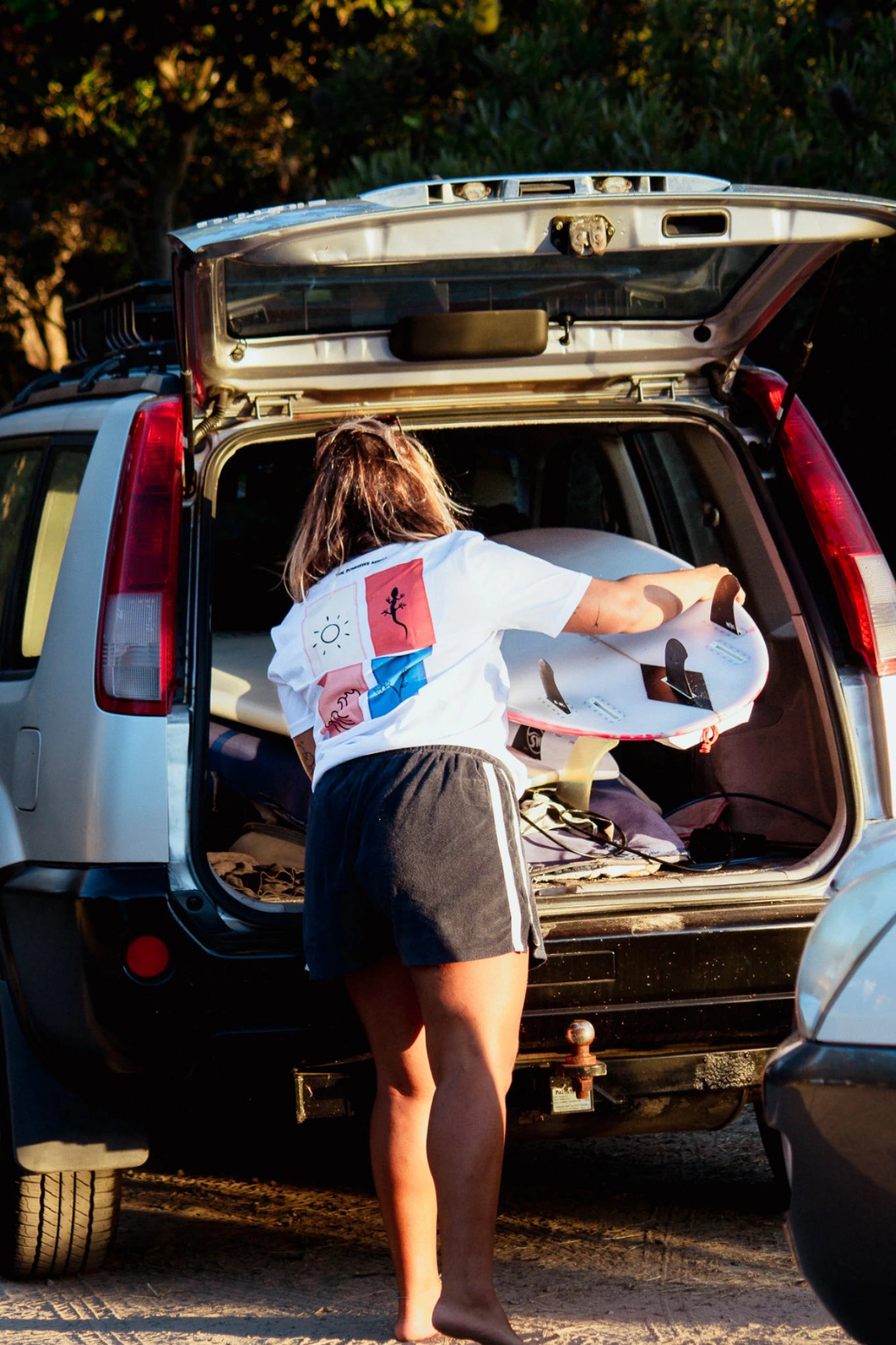 Frau lädt Surfboard aus Auto und trägt Rebel Elements Light T-Shirt in Weiß mit Backprint