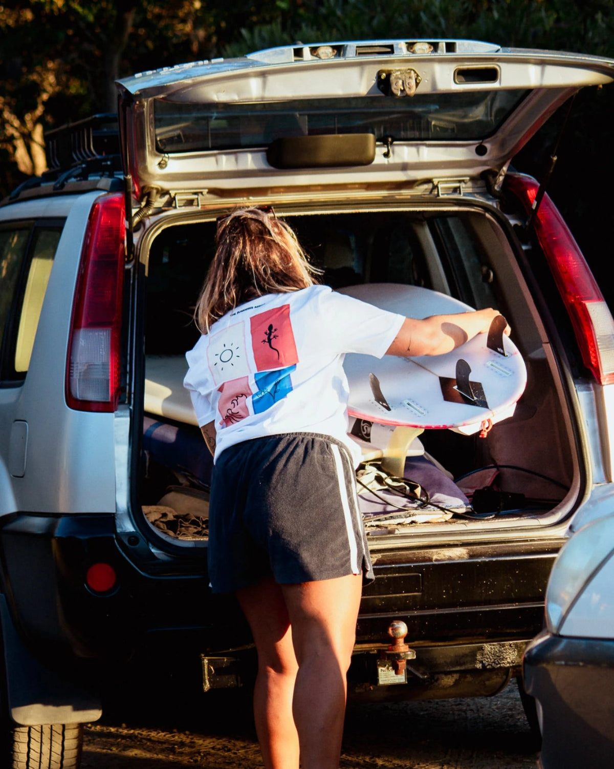 Frau lädt Surfboard aus Auto und trägt Rebel Elements Light T-Shirt in Weiß mit Backprint