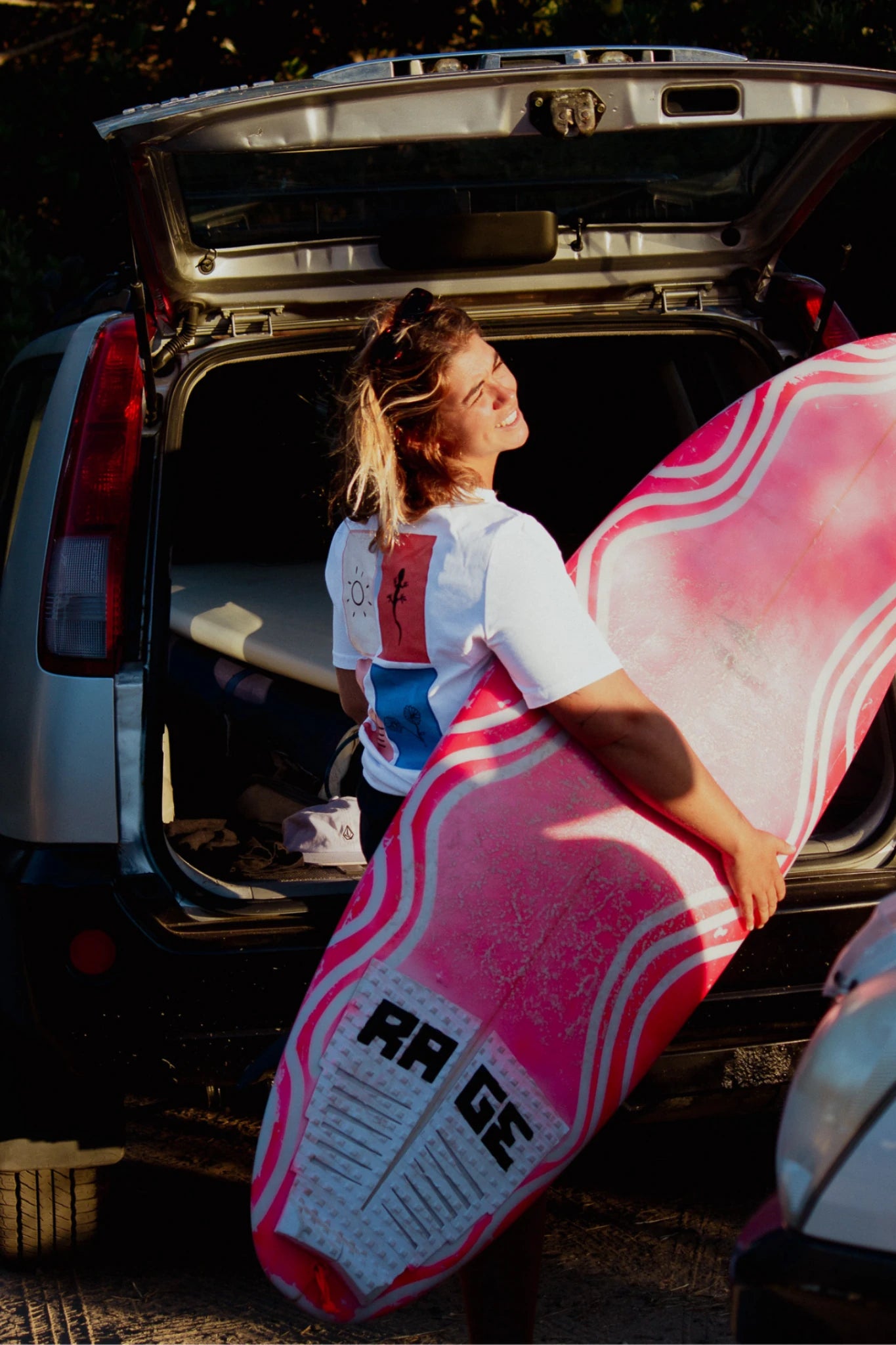 Frau mit pinkem Surfboard im Sonnenlicht trägt weißes Rebel Elements T-Shirt mit Grafik